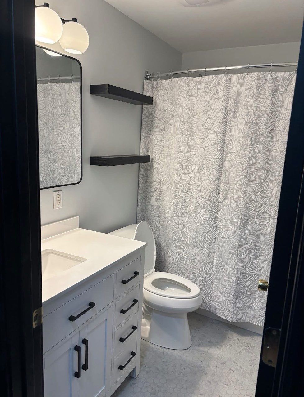 Small bathroom with white vanity, toilet, floating shelves, and gray shower curtain.