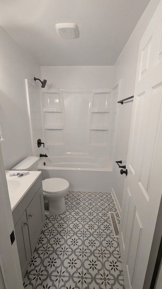 White bathroom with tub-shower, toilet, vanity, and patterned tile floor.