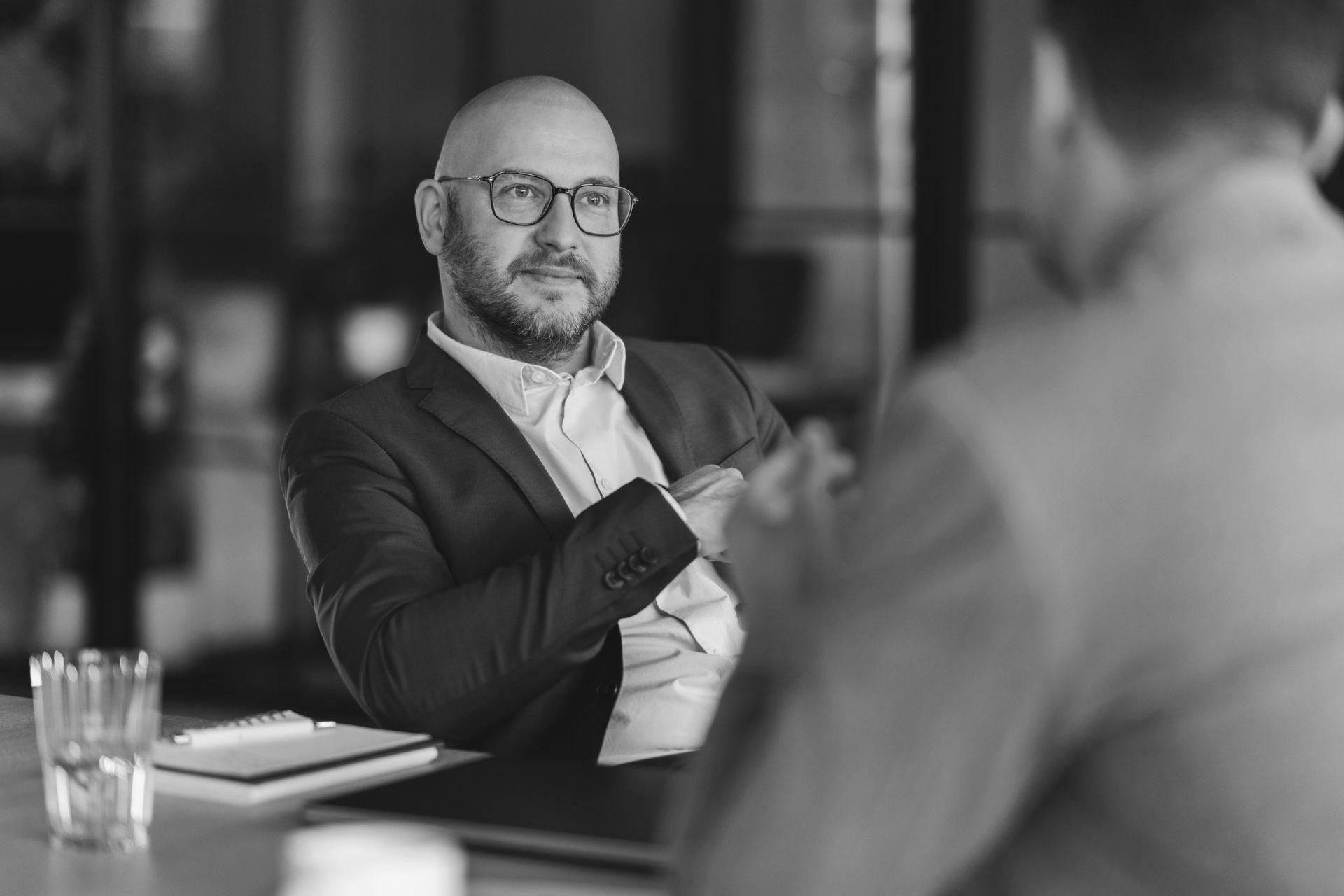 Man in a suit and glasses speaking at a table in a meeting room, with a laptop and glass on the desk.