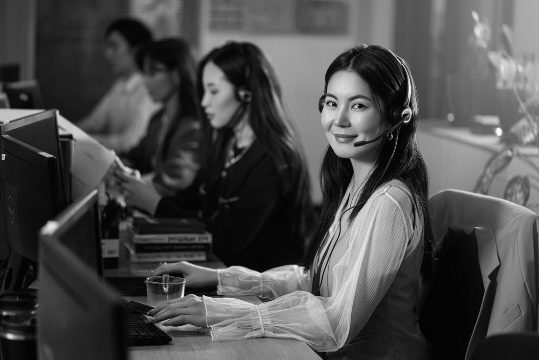 Woman smiling at a computer in a busy office, with coworkers in the background.