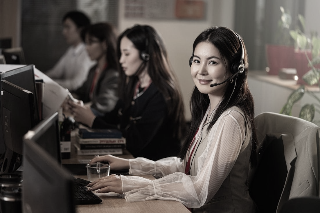Woman smiling at a desk in an office call center, wearing a headset and typing on a computer.
