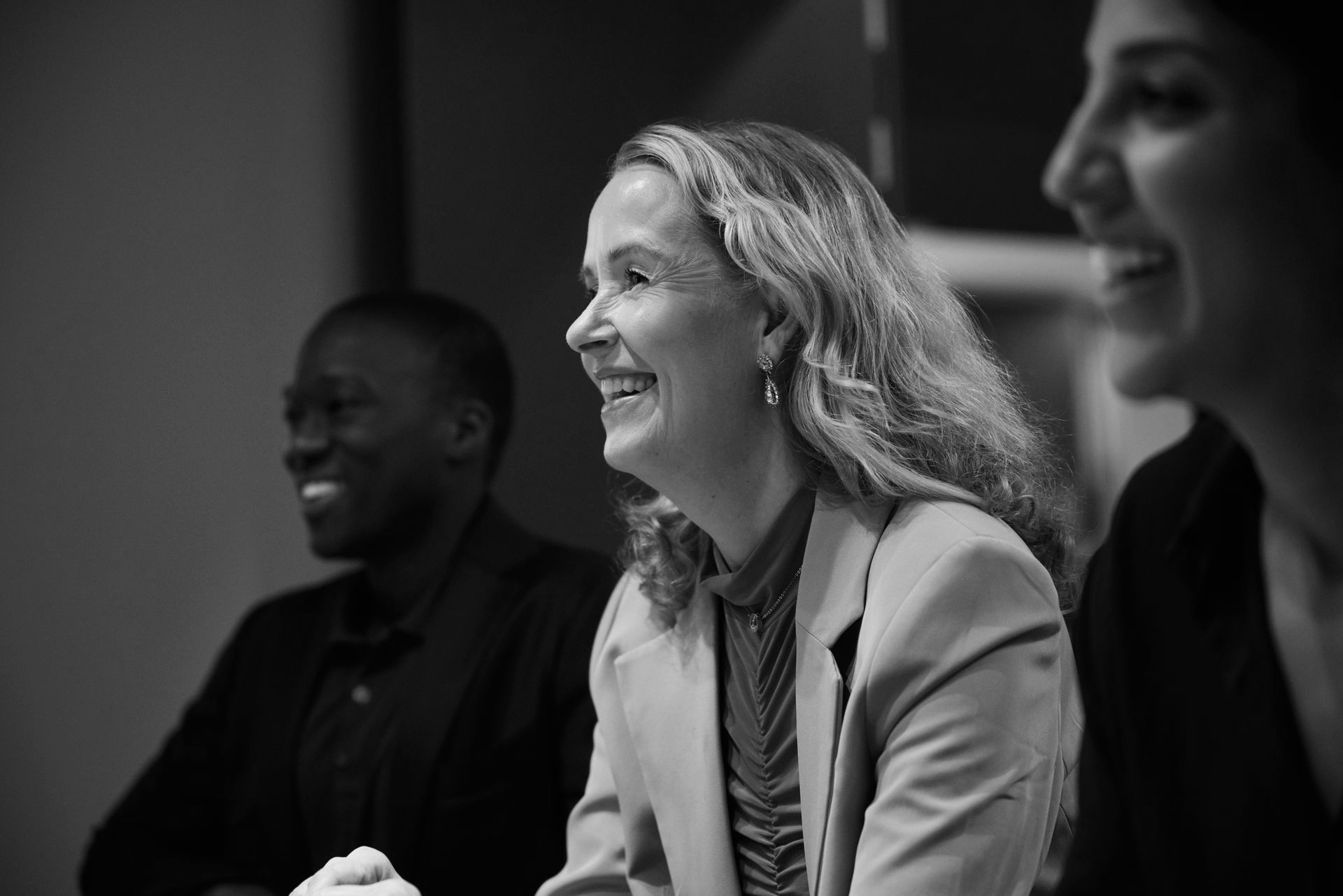 Three people smiling in a meeting, with a woman in a light blazer in focus.