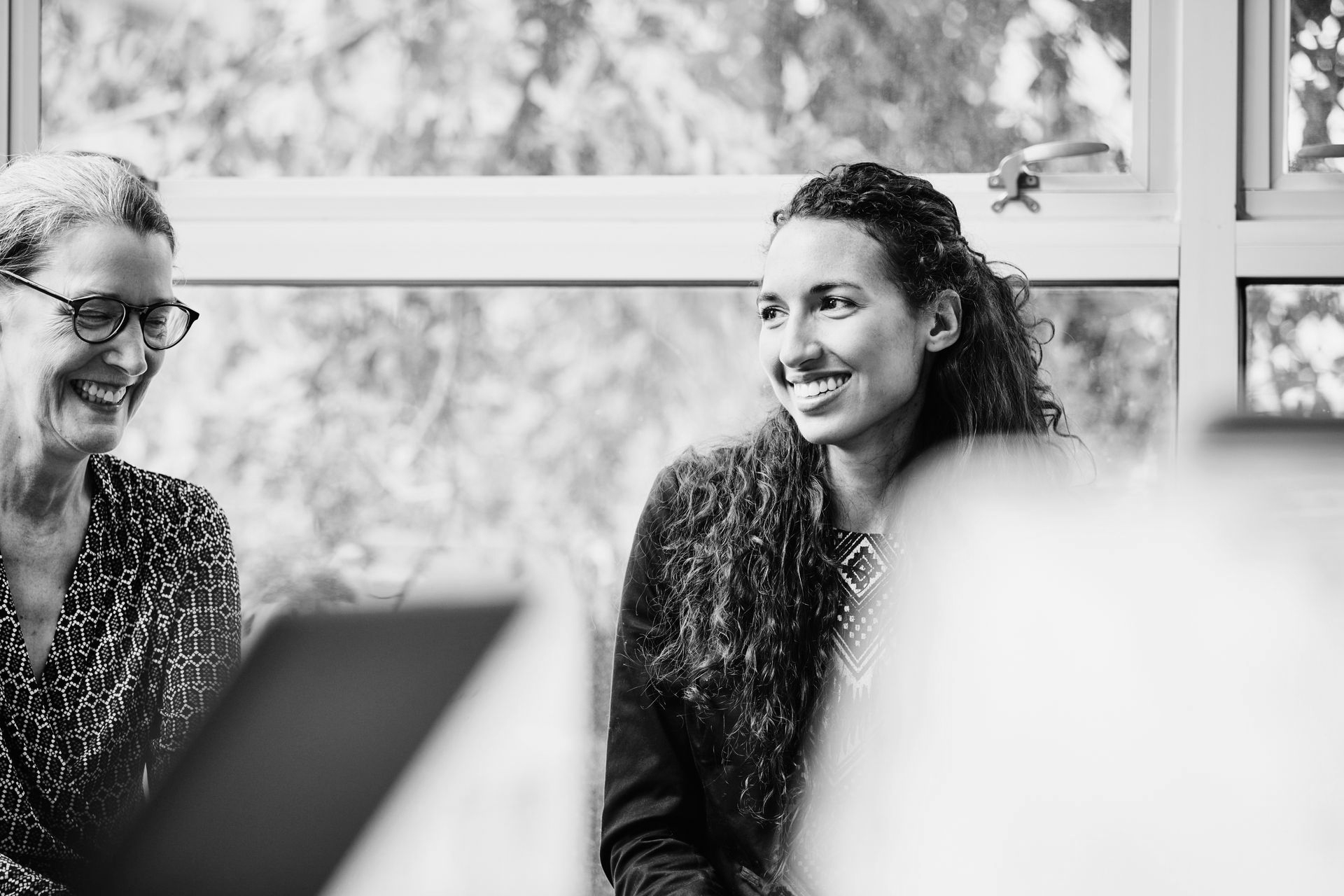 Two people smiling and talking in a bright office, seen in black and white.