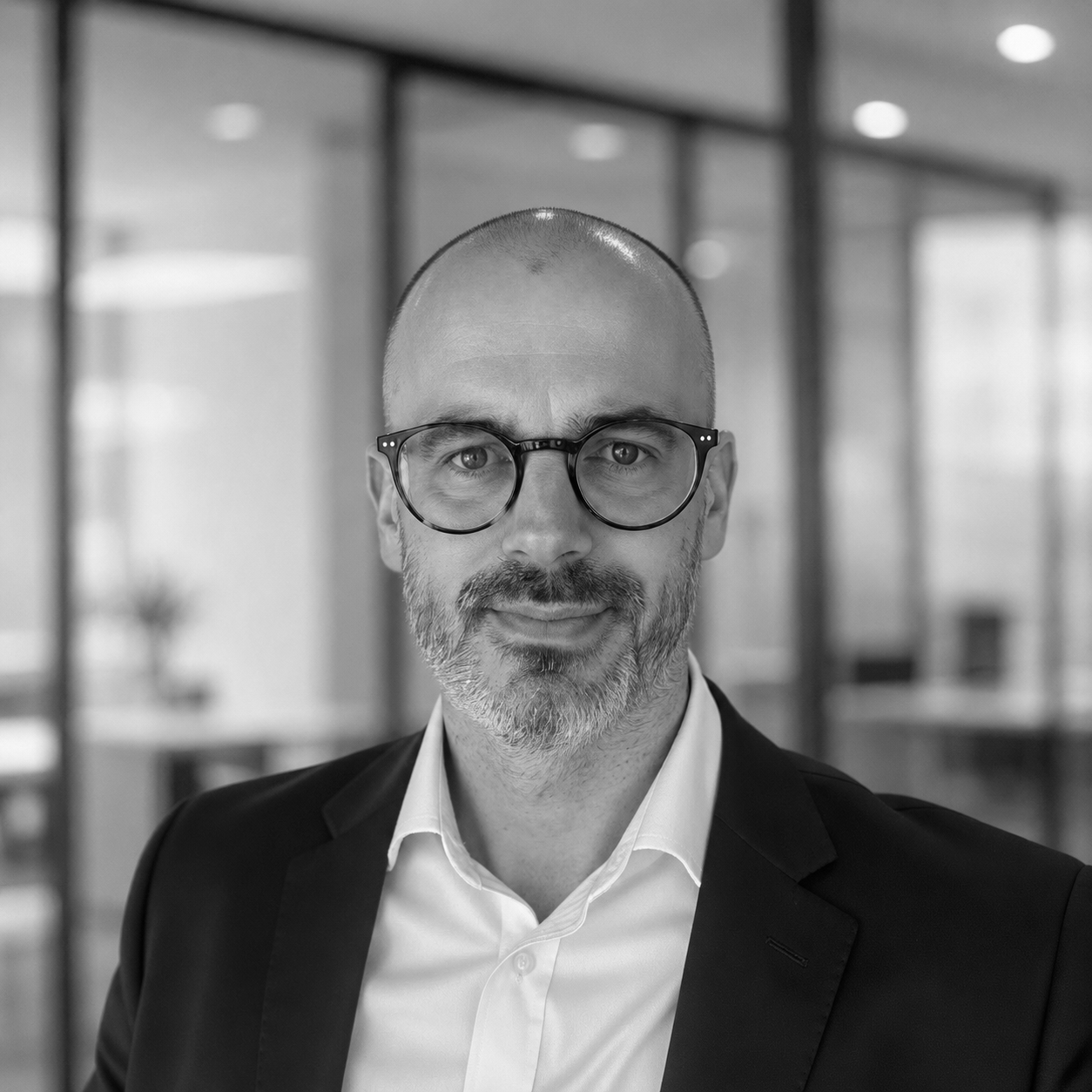 Man in glasses and blazer standing in a modern office, looking at the camera.