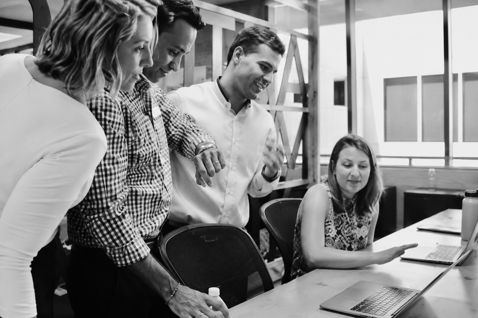 Four coworkers collaborate around a laptop in a modern office, smiling and focused.