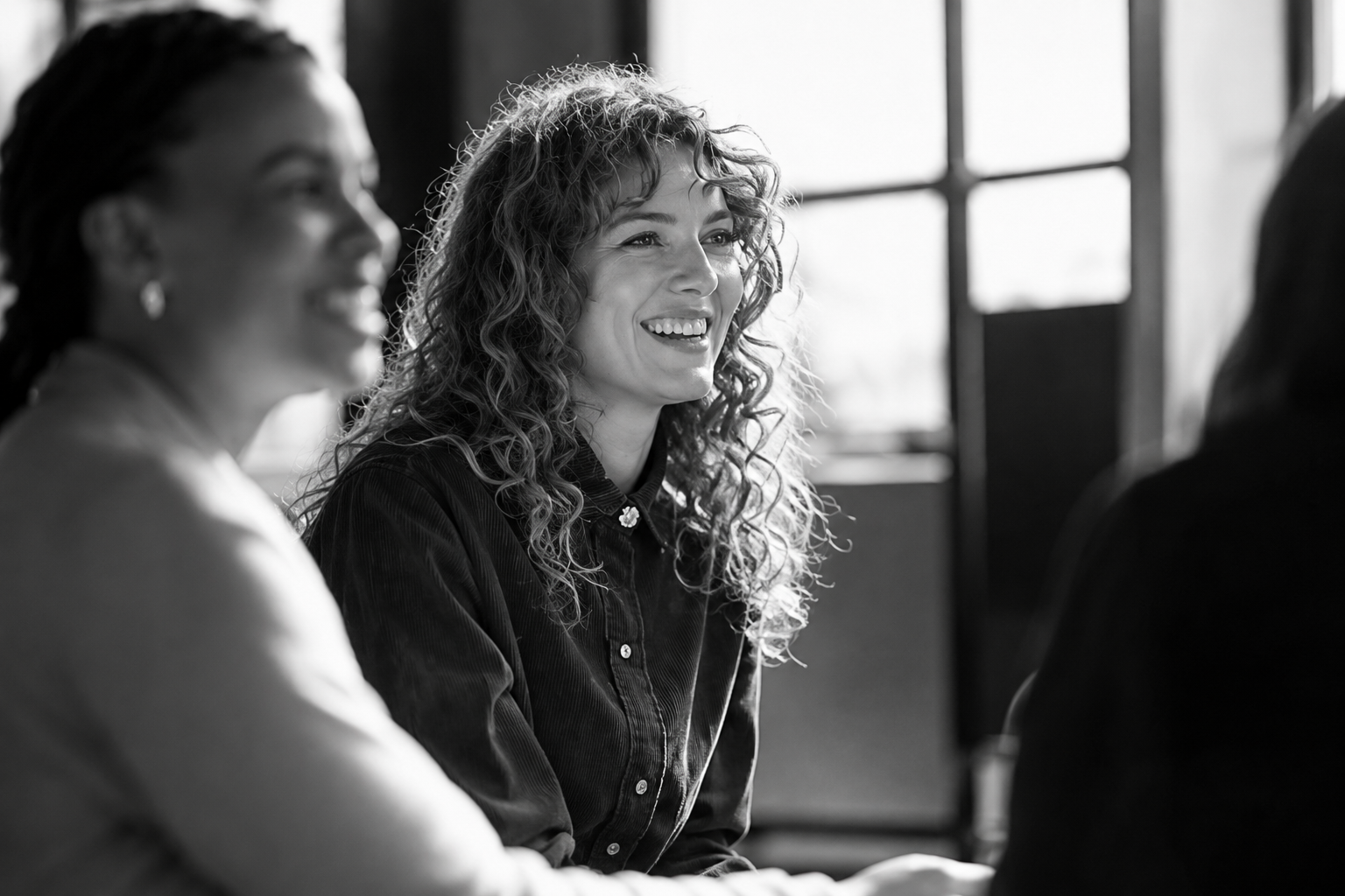 Two people smiling in a sunlit meeting room, with a blurred third person in the foreground
