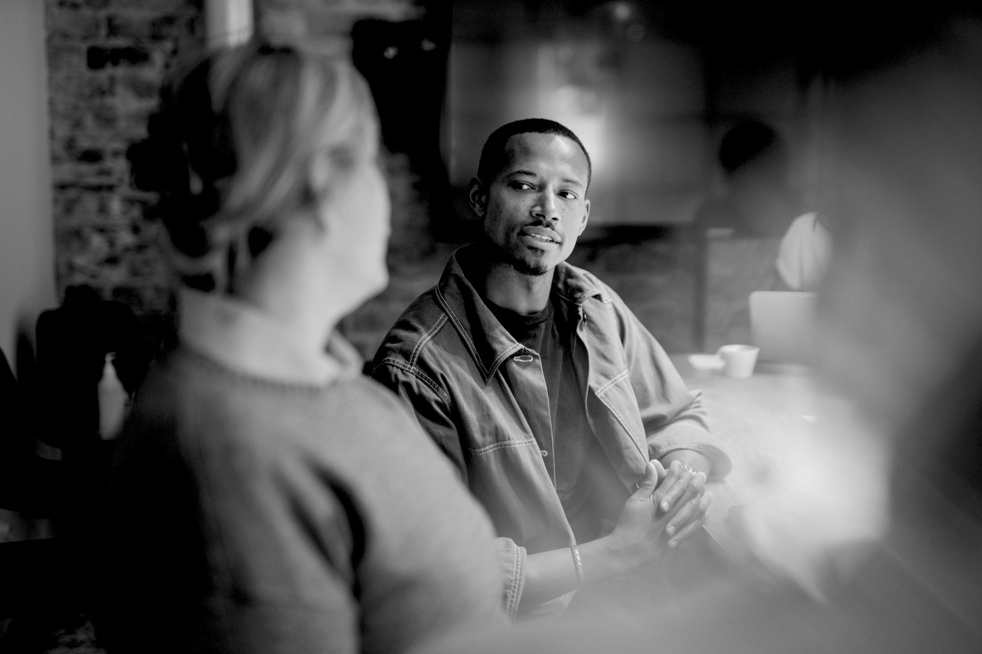 Two people sit in a dim café, one in focus speaking while the other listens in the foreground.