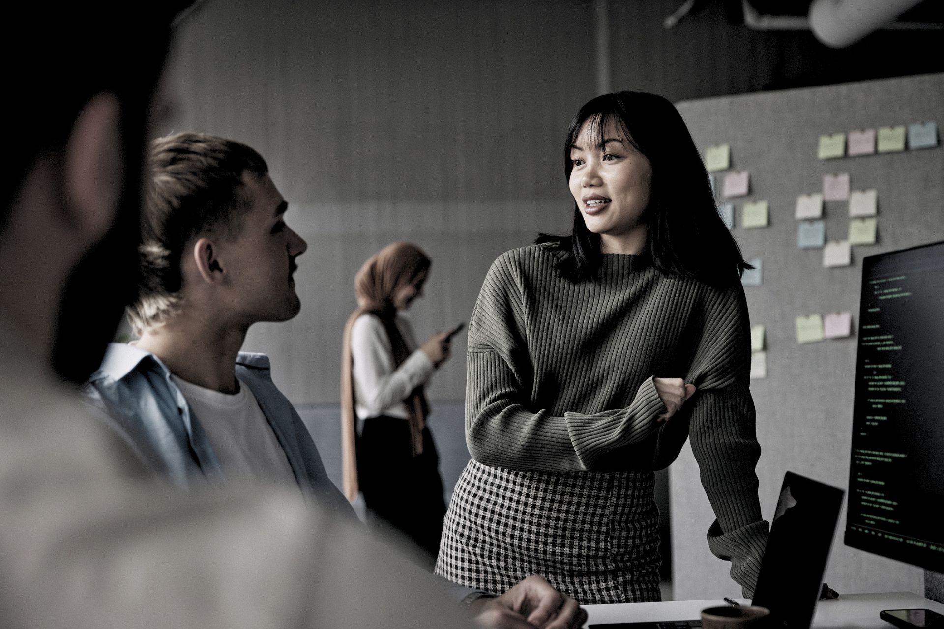 Colleagues talking in an office, with one woman standing by a computer monitor and sticky notes on the wall.