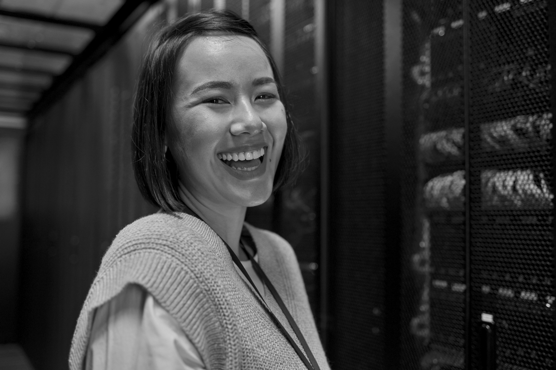Smiling person standing in a hallway beside server racks, wearing a light sweater.