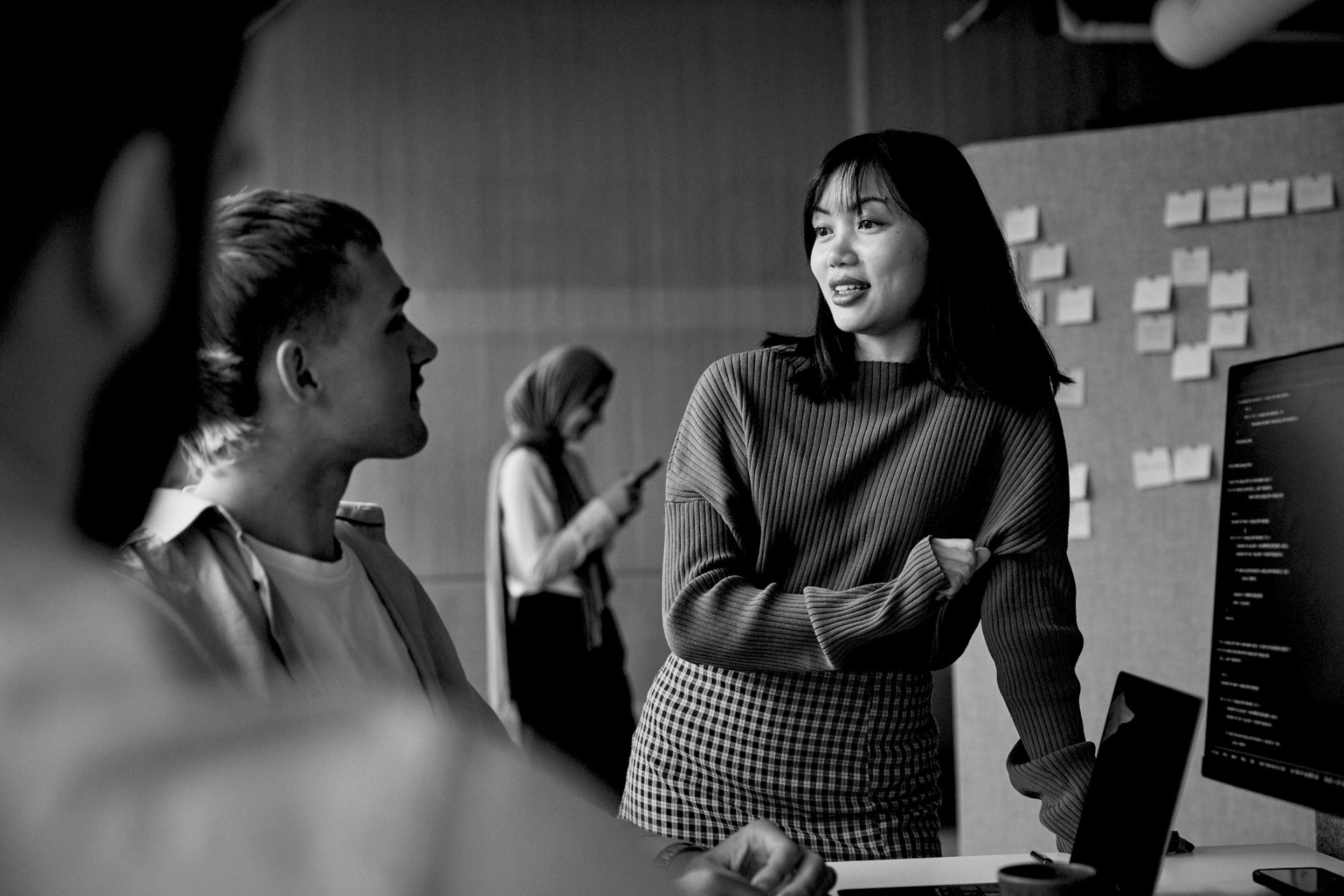 Two coworkers talking in an office, with one standing by a computer monitor and notes on the wall.