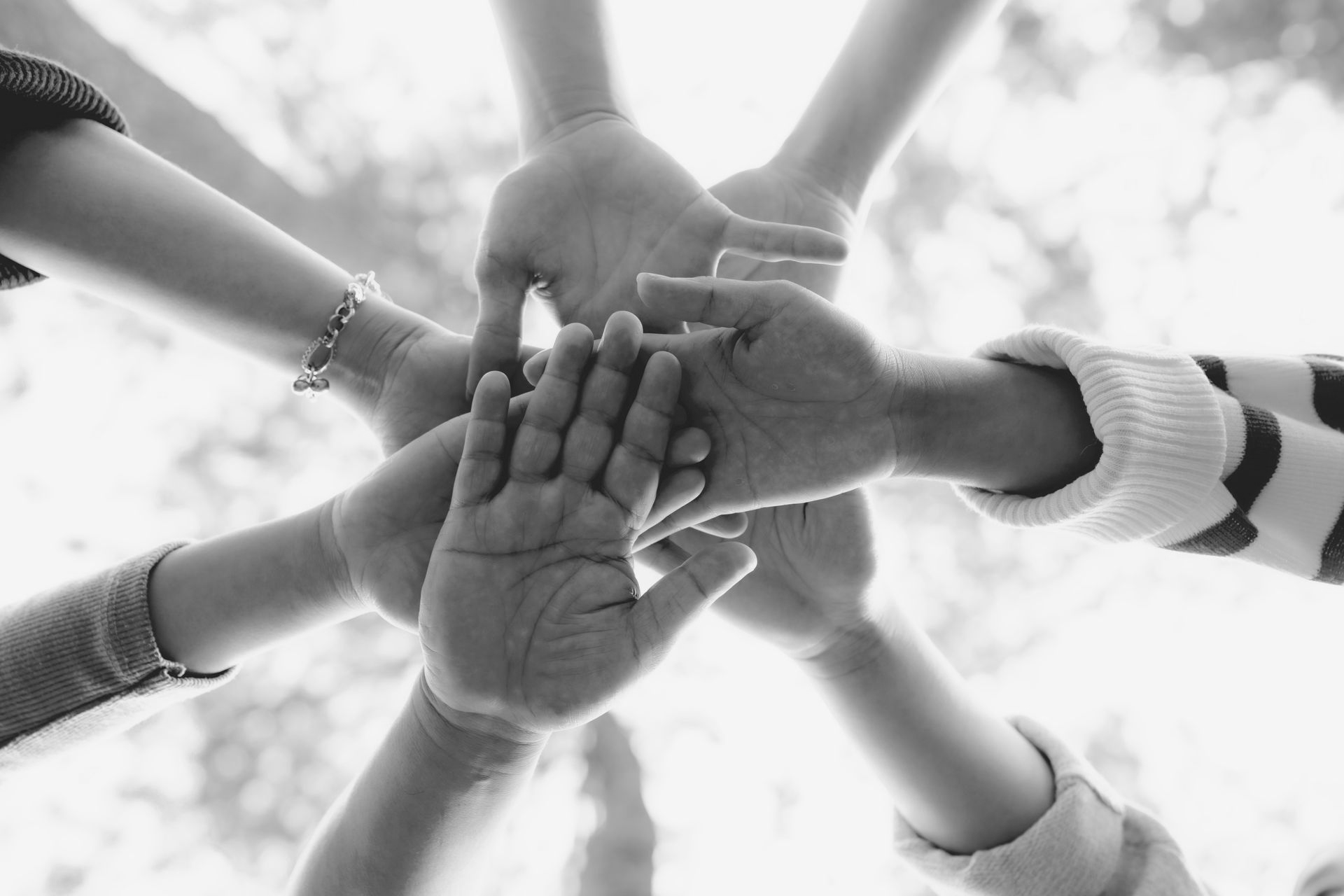 Several hands reaching inward in a circle against a bright sky, viewed from below