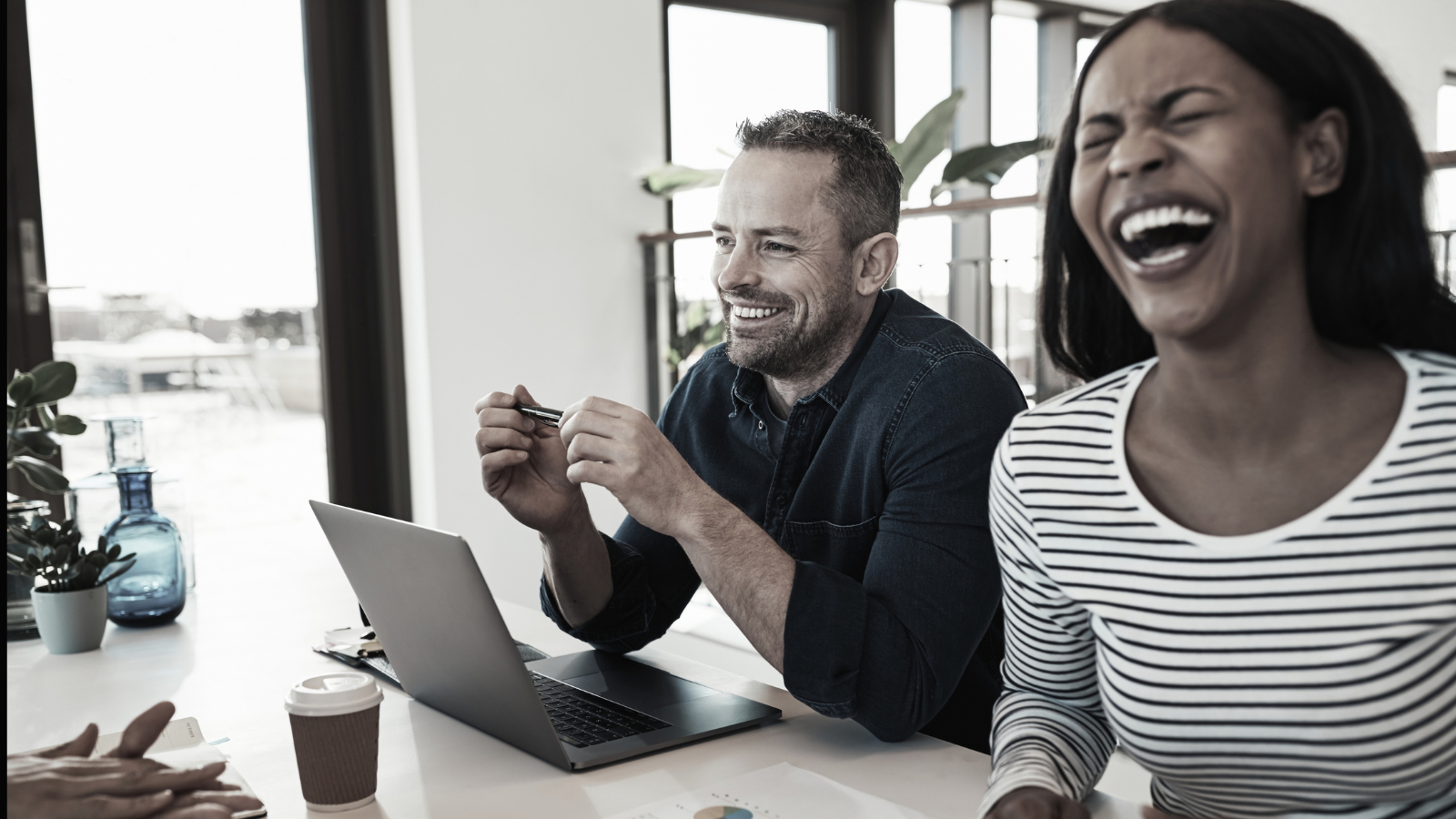 Two people laughing at a desk with a laptop in a bright office