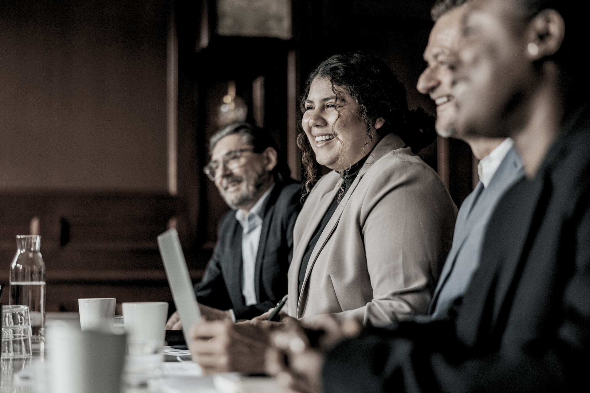 Three people in business attire smiling during a meeting at a conference table with a laptop and water bottle.