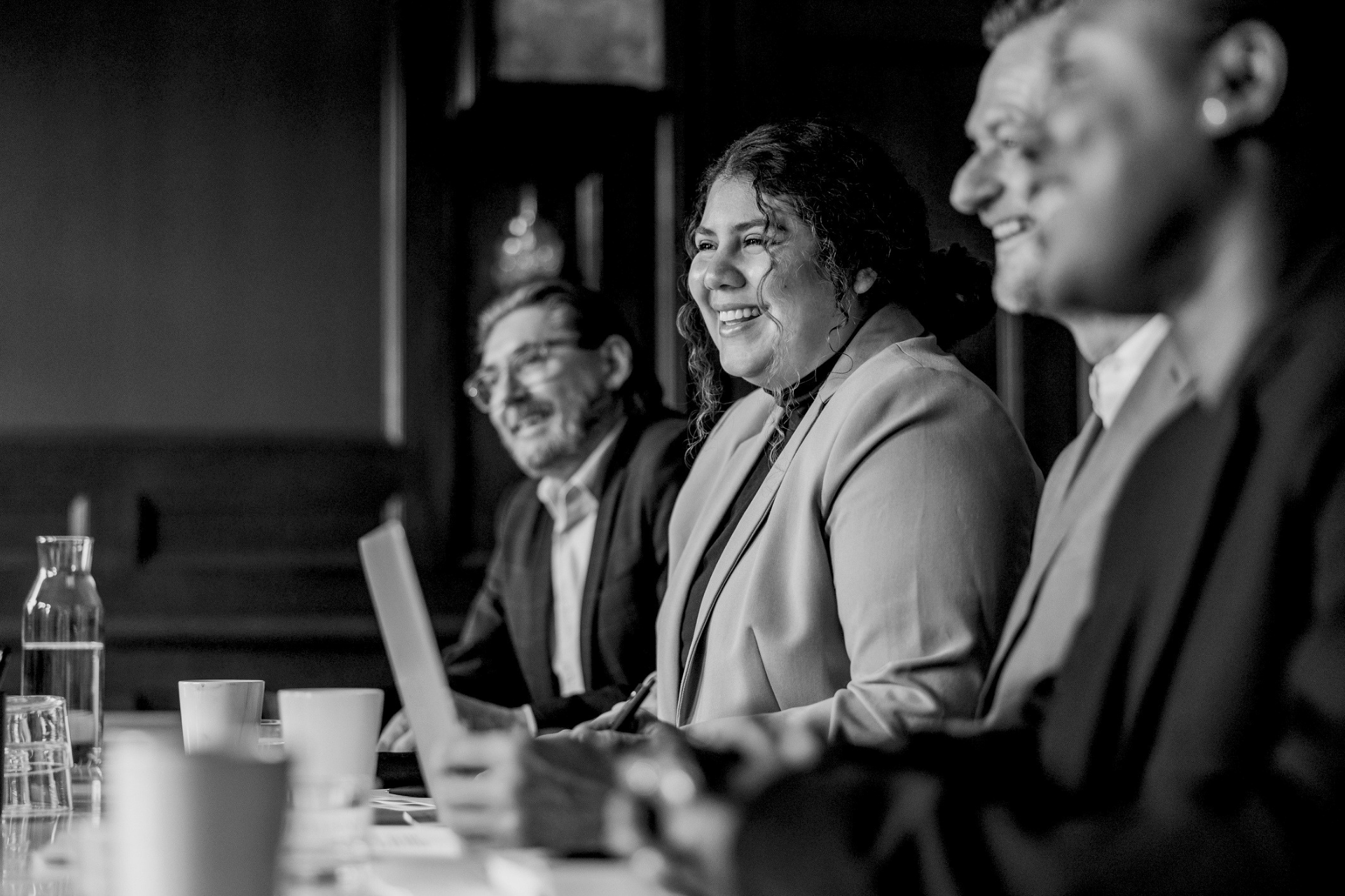 Three people seated at a meeting table, smiling and listening in a black-and-white office setting.