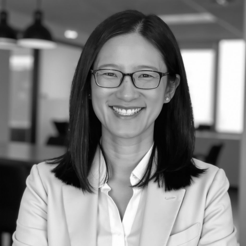 Smiling person with glasses in a light blazer, standing in an office setting.