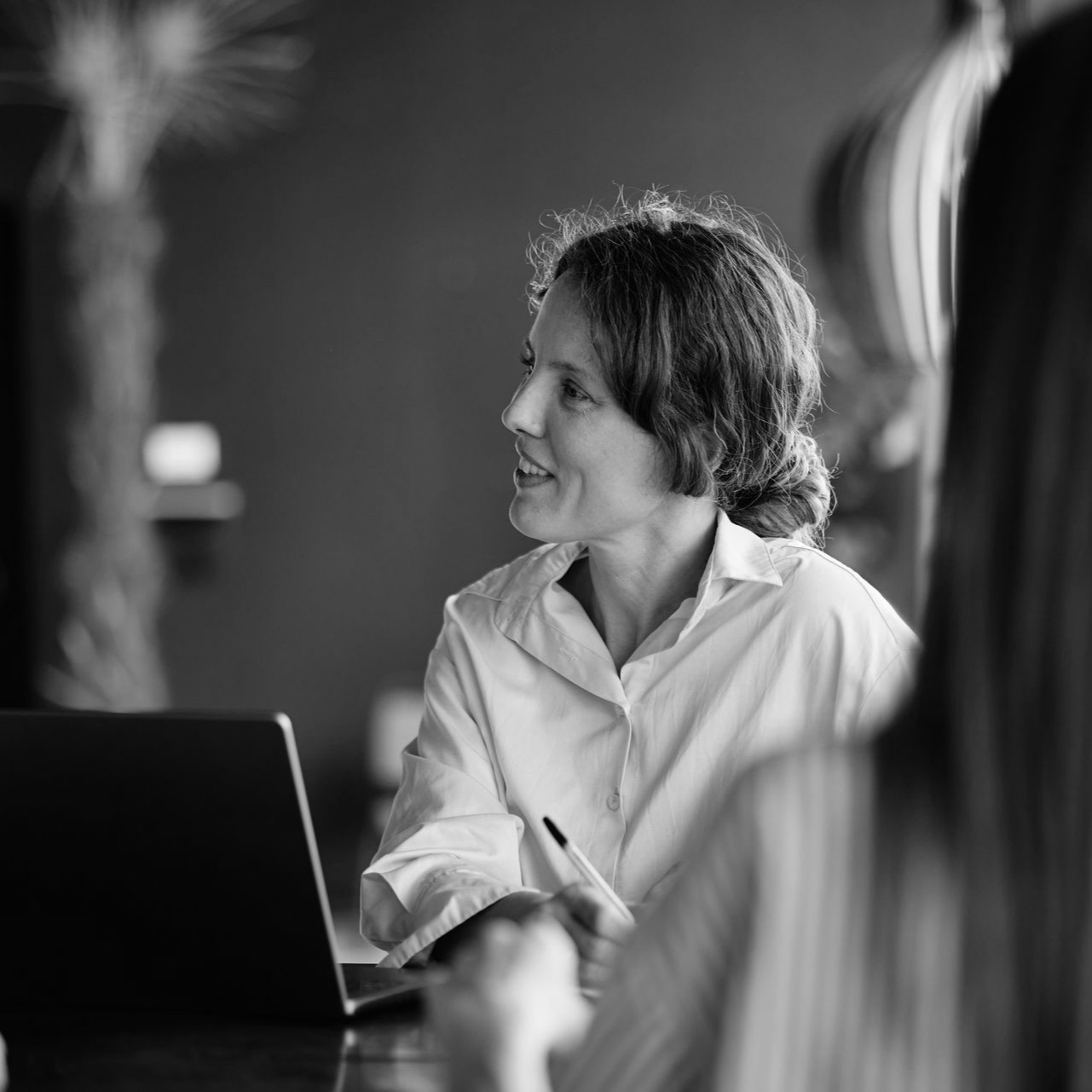 Woman in a white shirt writing beside a laptop in a meeting room, smiling and looking to the side