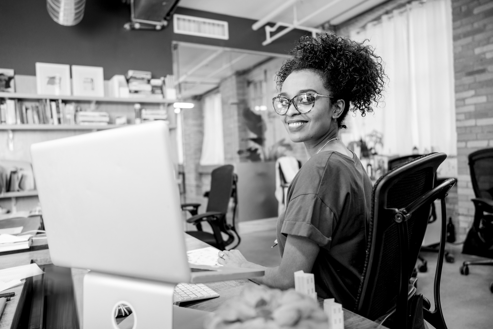 Woman smiling at a desk with a computer in a dim office, coworkers blurred in the background