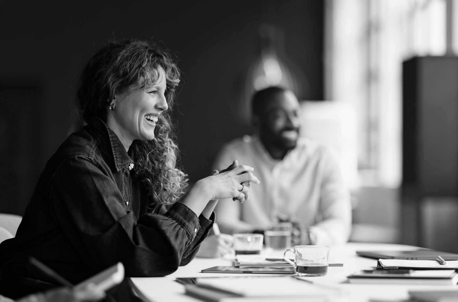 People in a meeting, smiling and talking around a conference table with papers and coffee cups.