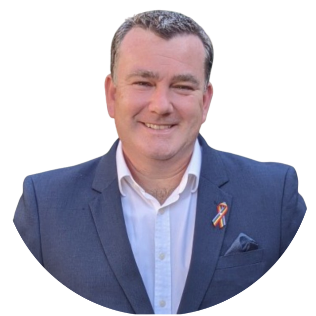 Smiling man in a blue blazer and white shirt with an orange ribbon pin, against a plain white background