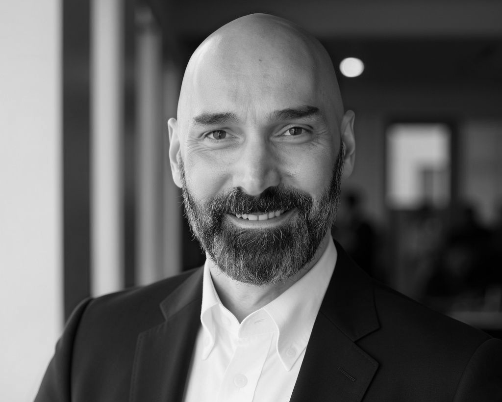 Smiling bald man in a dark suit and white shirt, posing indoors in black and white.