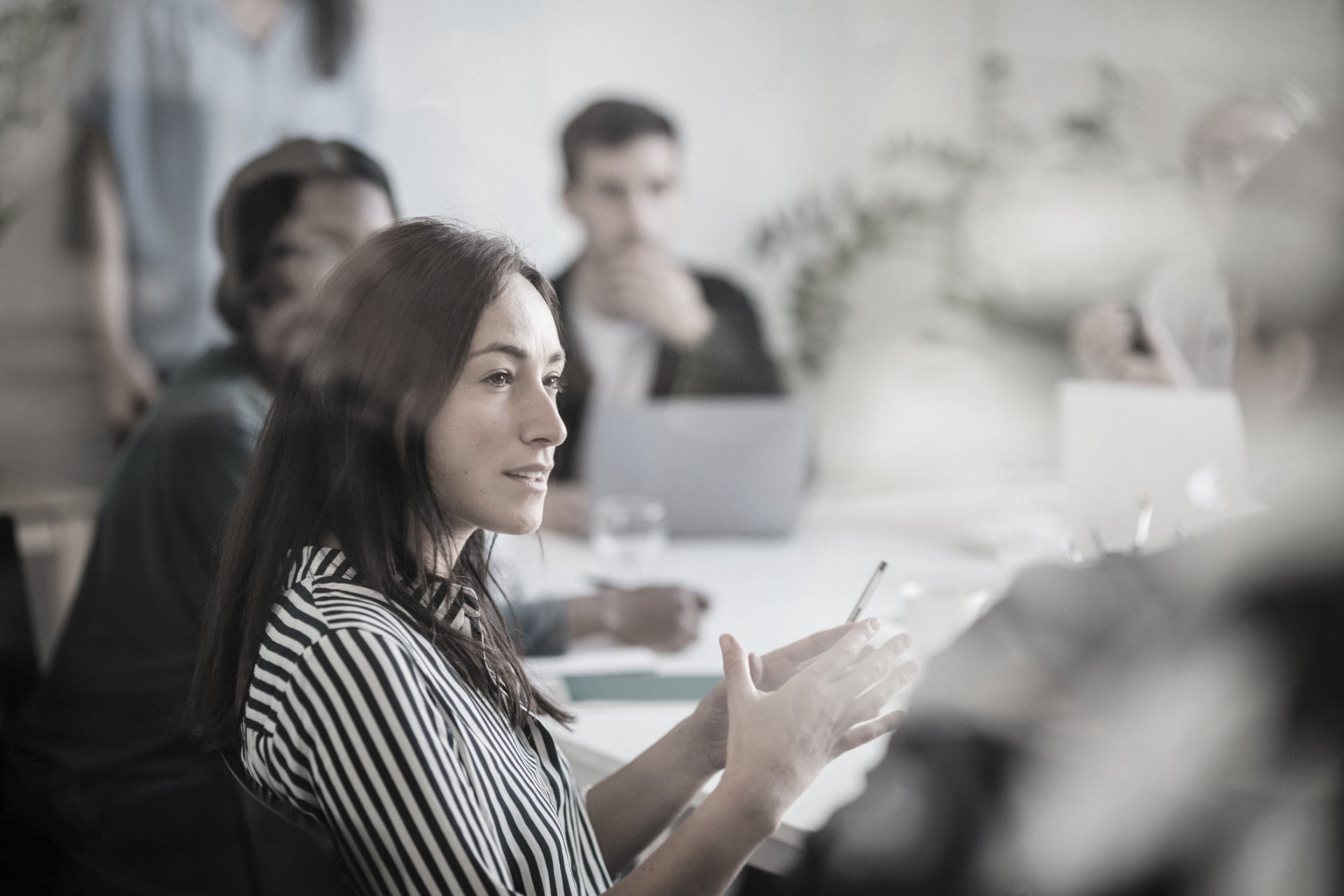 Woman speaking in a meeting room, gesturing while seated at a table with colleagues and laptops.