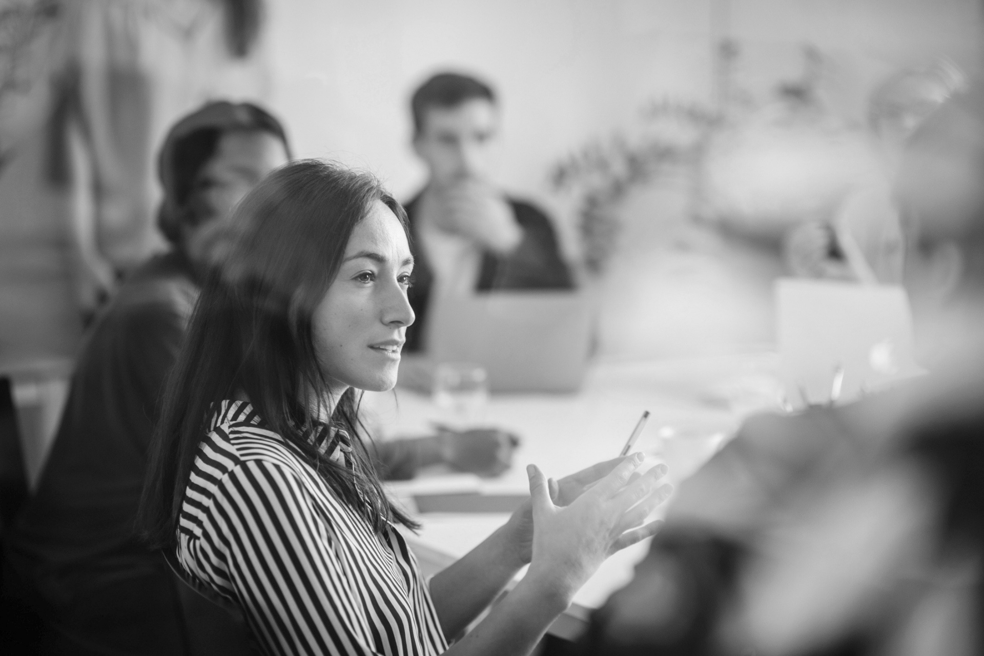 Woman speaking at a meeting table, gesturing with her hands in a blurred office setting.