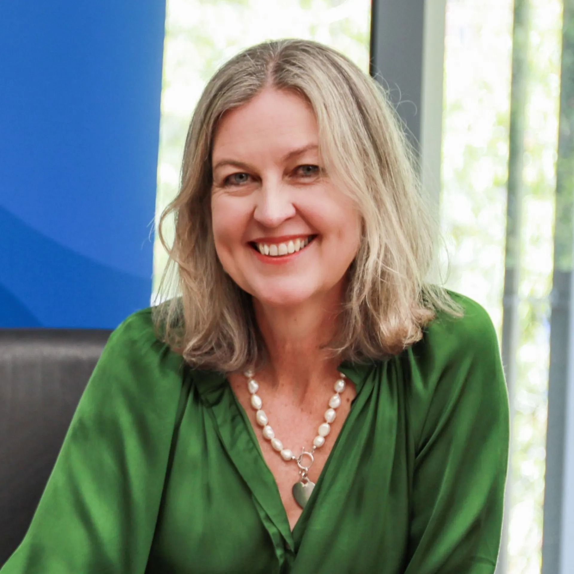 Smiling woman in green blouse and pearl necklace seated indoors against a blue and white background