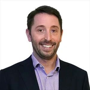 Smiling man in a dark blazer and light purple shirt against a white background