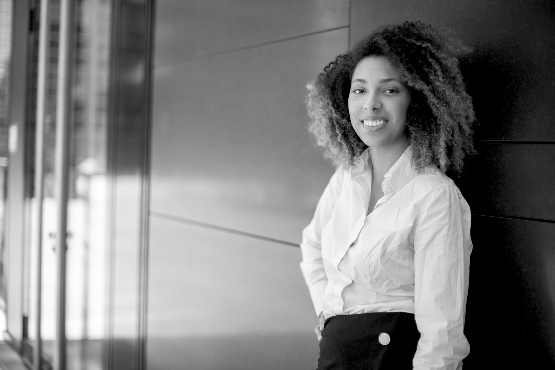 Smiling woman in a white blouse leaning against a wall in a modern hallway.
