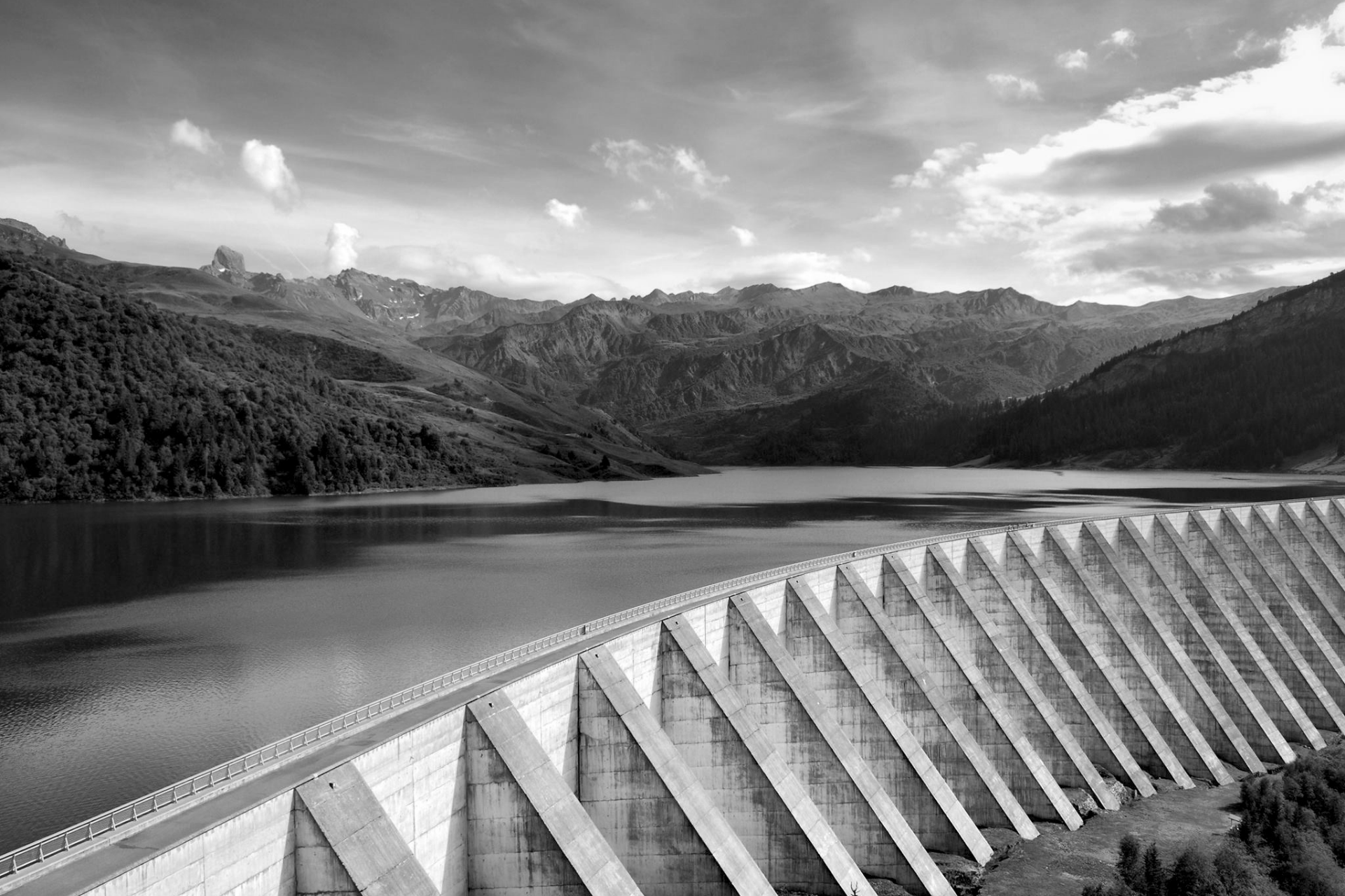 Black-and-white view of a dam beside a calm lake, with mountains and clouds in the background