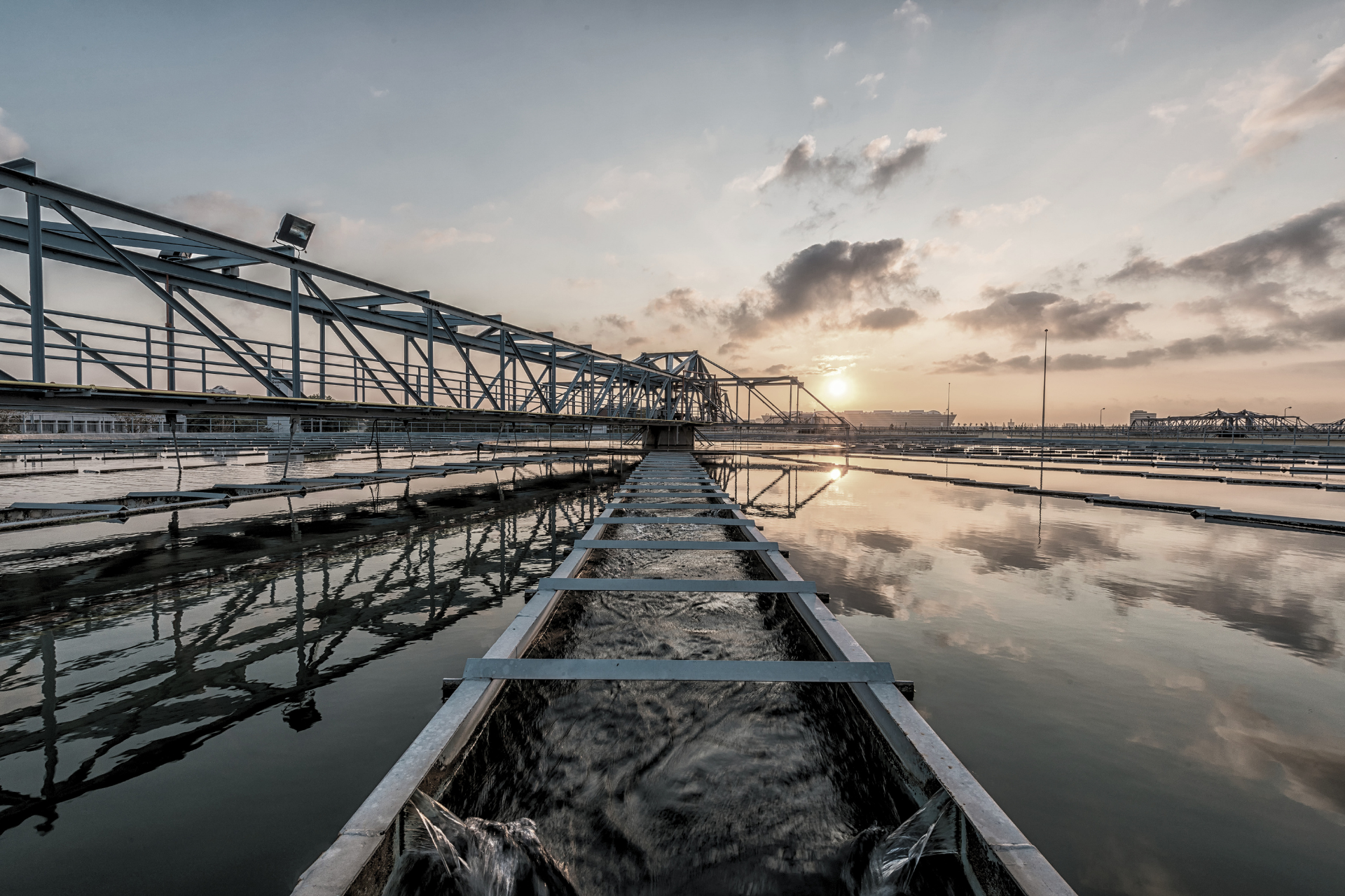Industrial walkway over reflective water at sunset with metal structures and cloudy sky