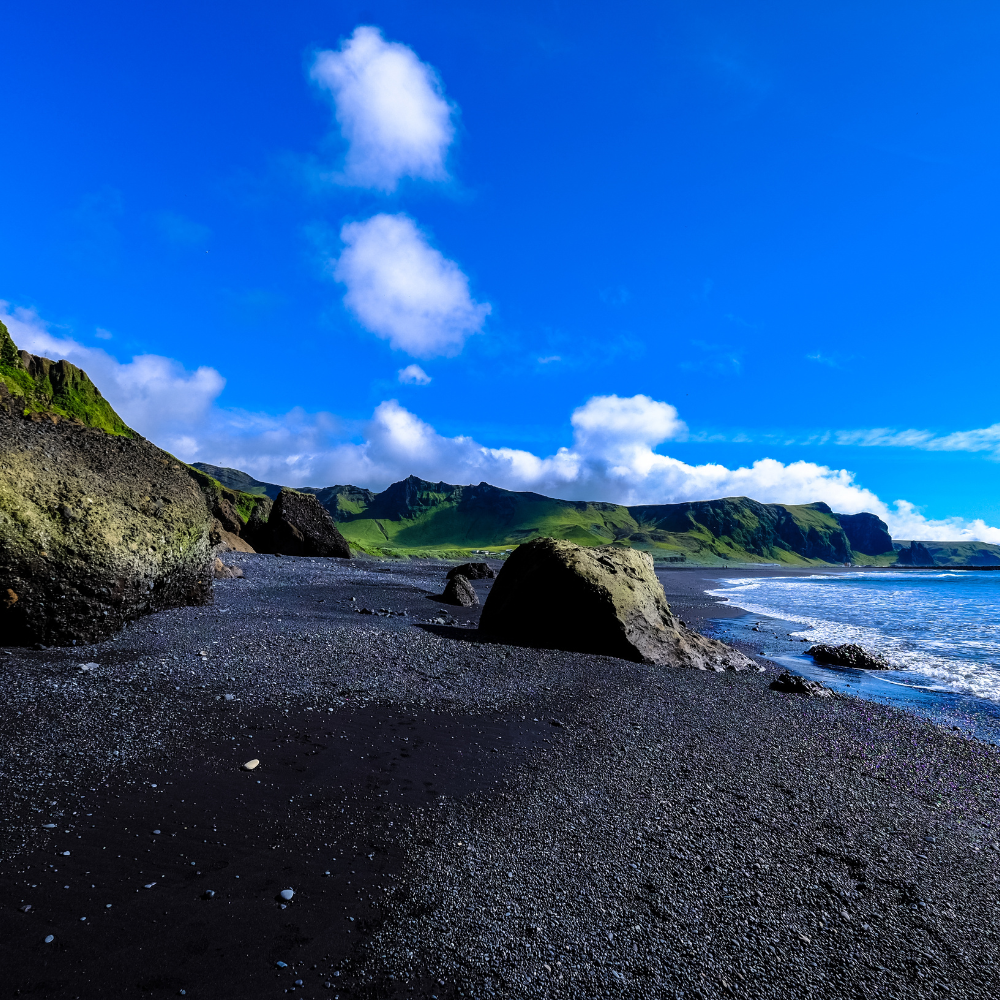 A black sand beach with dark rocky formations under a vibrant blue sky with scattered white clouds and ocean waves.