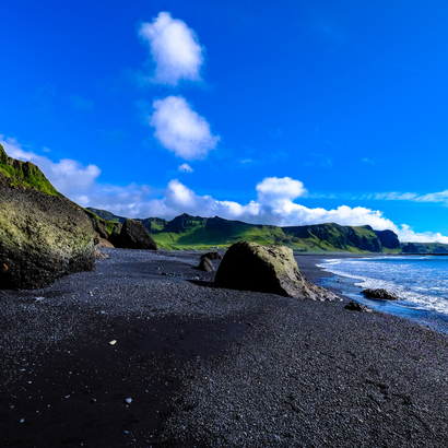 A black sand beach with dark rocky formations under a vibrant blue sky with scattered white clouds and ocean waves.