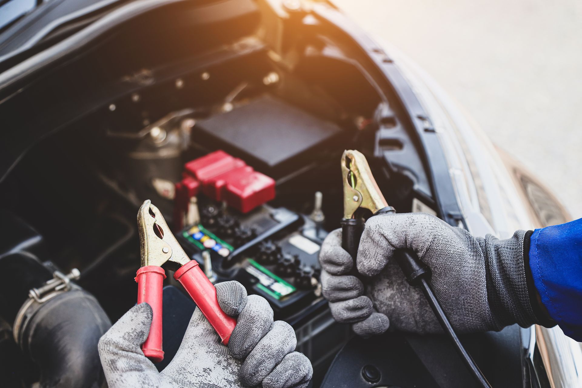 Hands wearing gloves connecting jumper cables to a car battery under the hood.