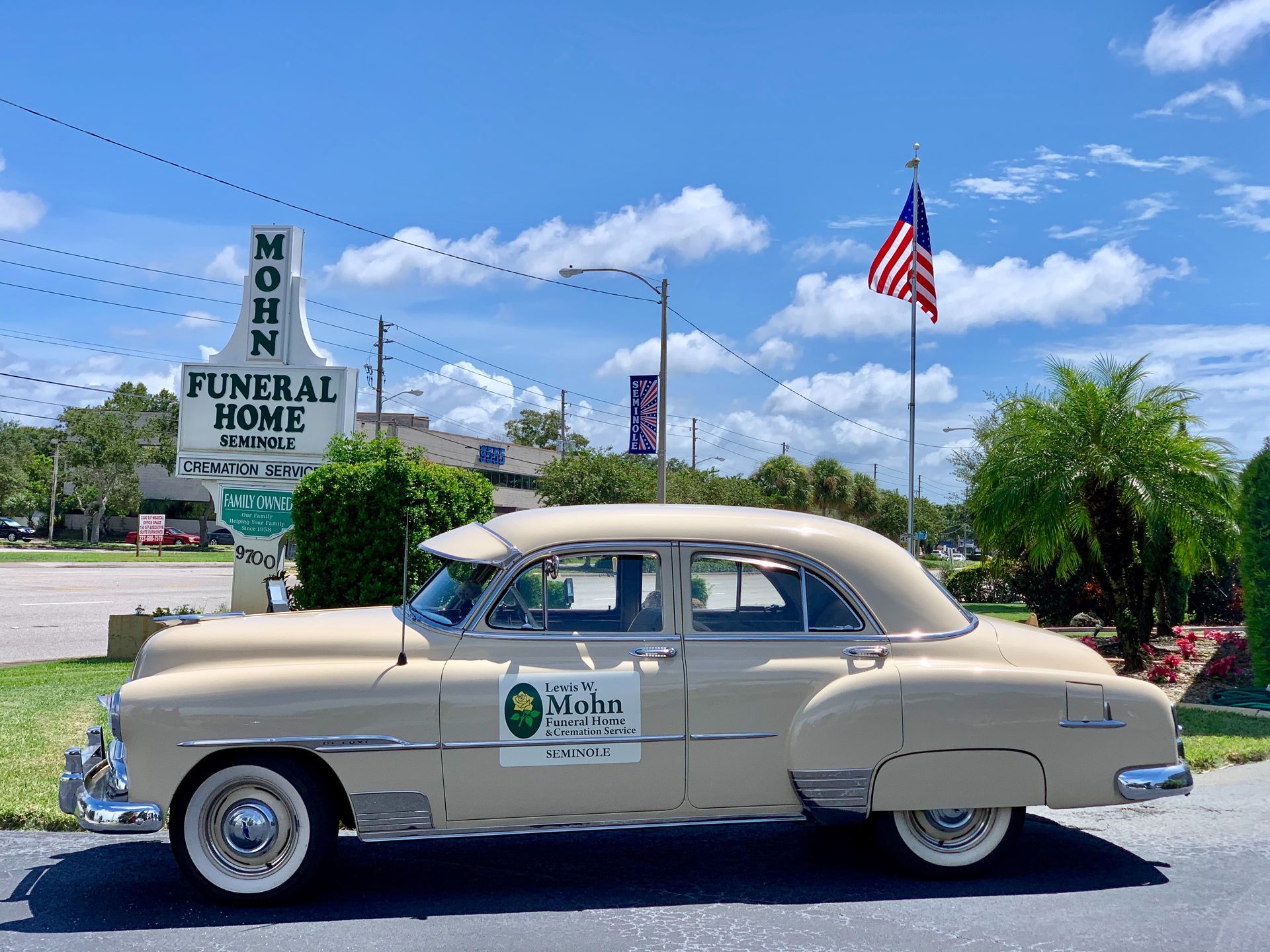 Vintage beige sedan parked outside a funeral home under a blue sky with an American flag.