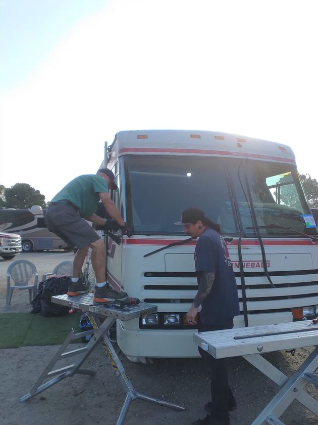 Two people working on a white RV outdoors beside folding tables at dusk