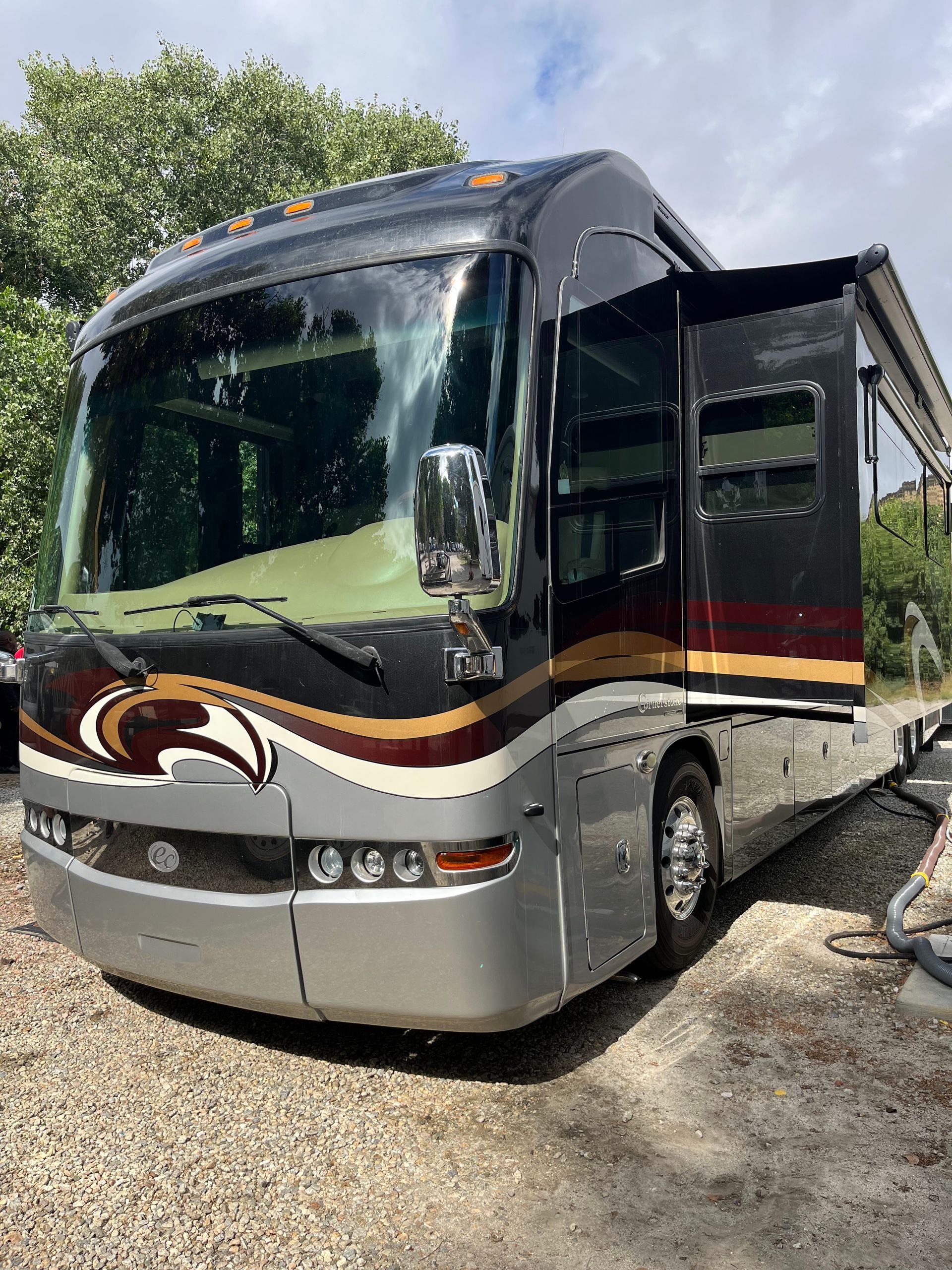 Silver tour bus with maroon and gold stripes parked outdoors, front door open in bright sunlight