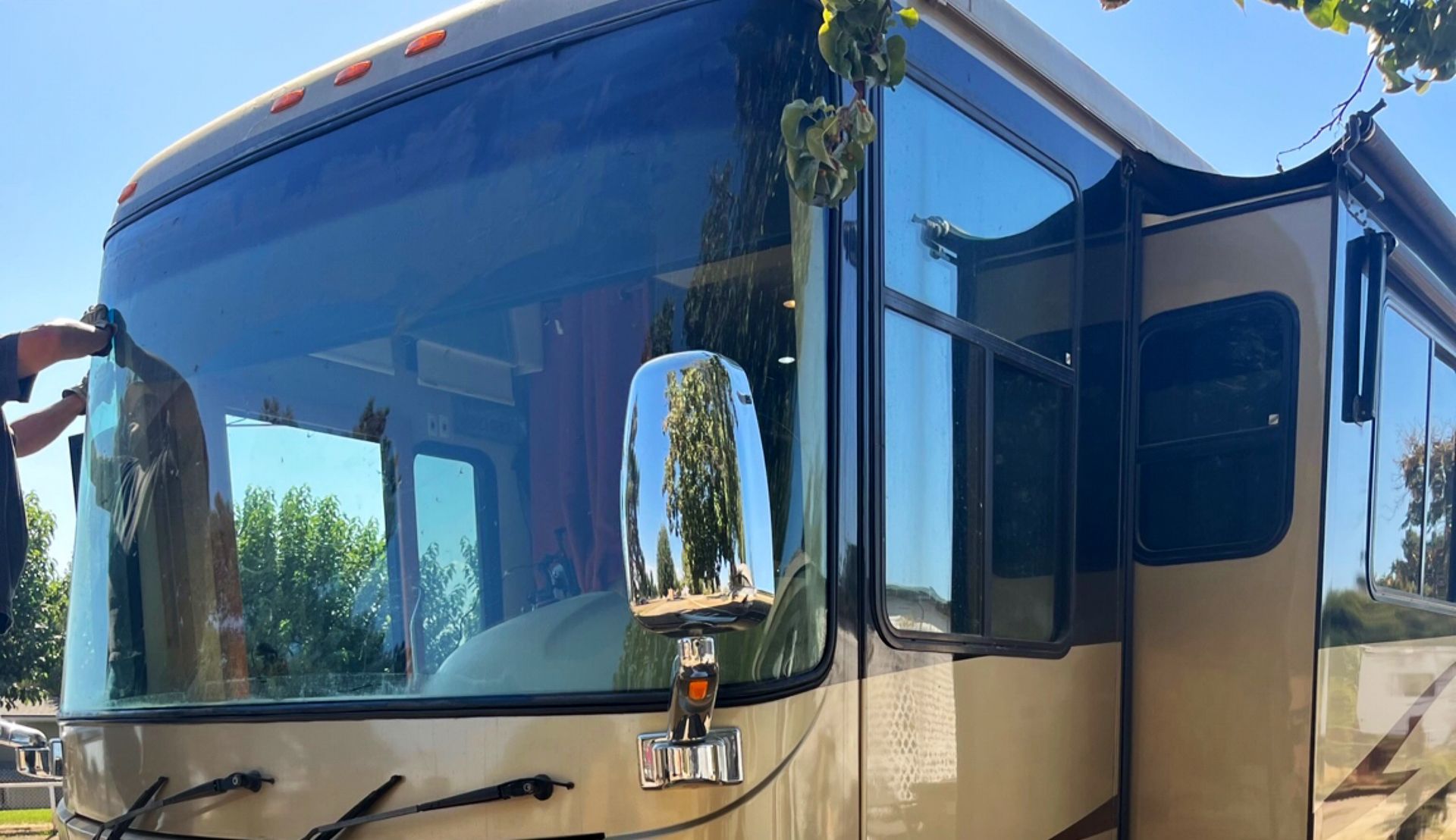 Large tan RV with dark tinted windows and open door parked outdoors in bright sun