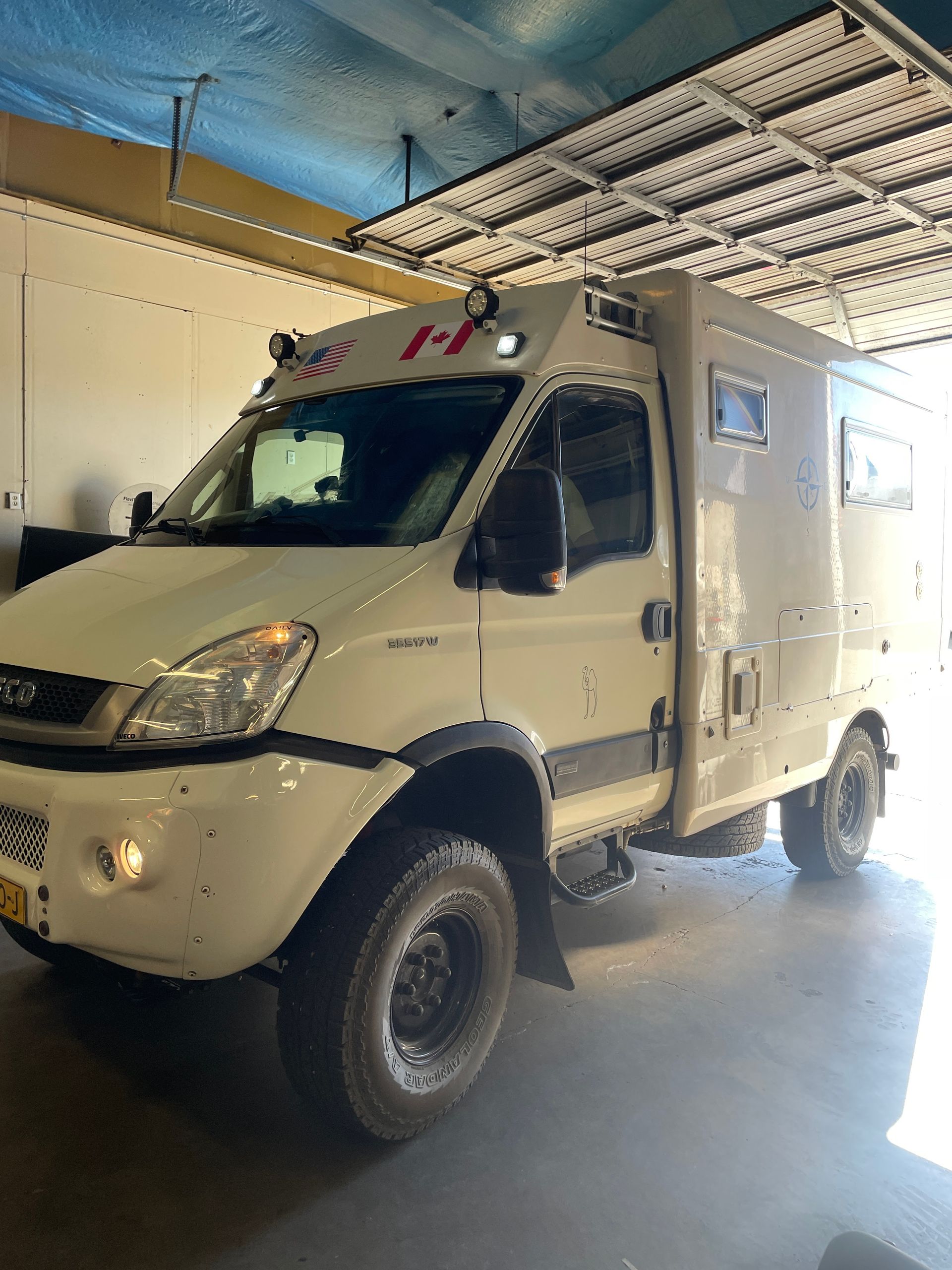 White armored van parked indoors under a blue ceiling, shown from the front-left side.