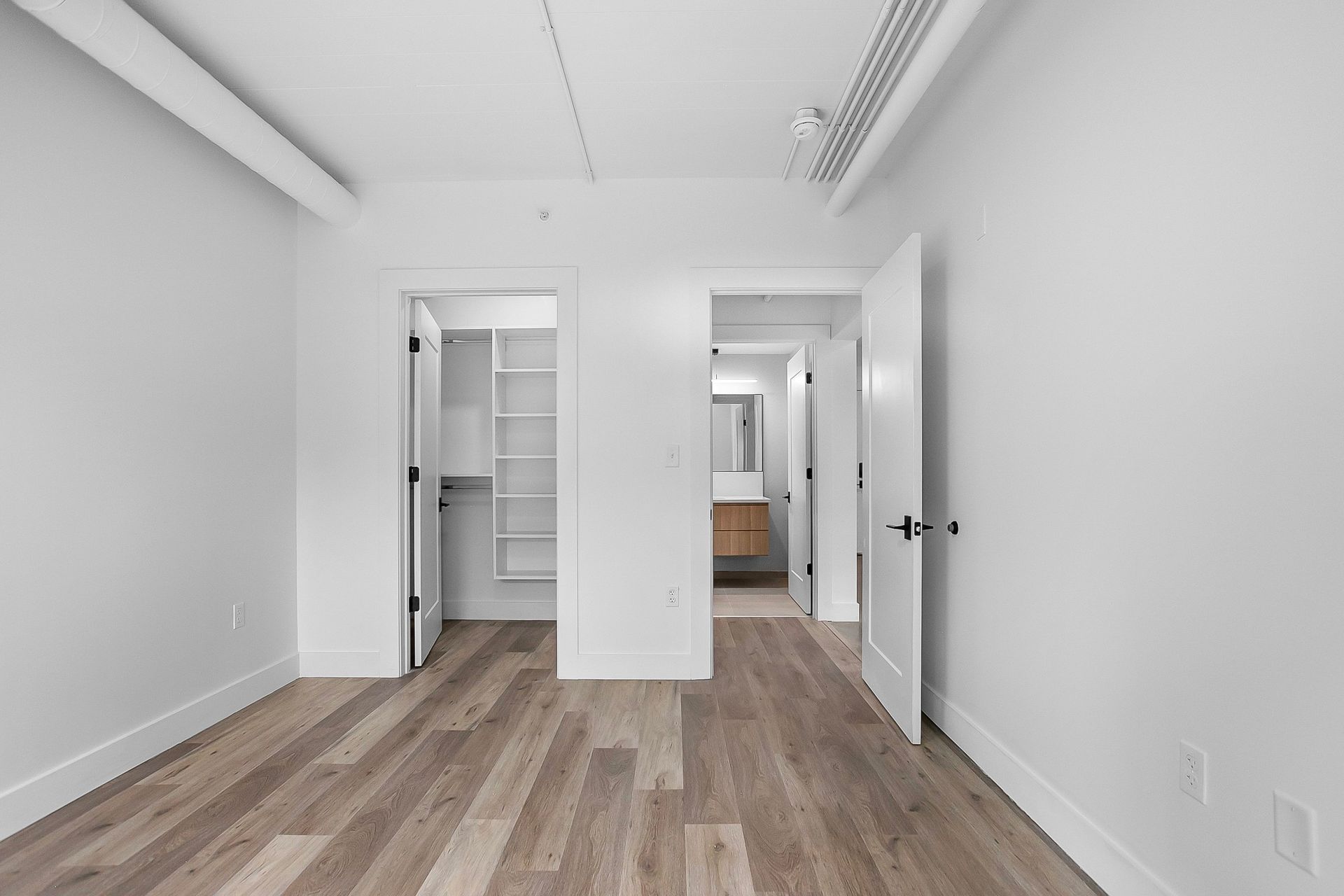 Empty white-walled room with wood-look flooring, two doorways, one leading to a closet and the other to a bathroom.