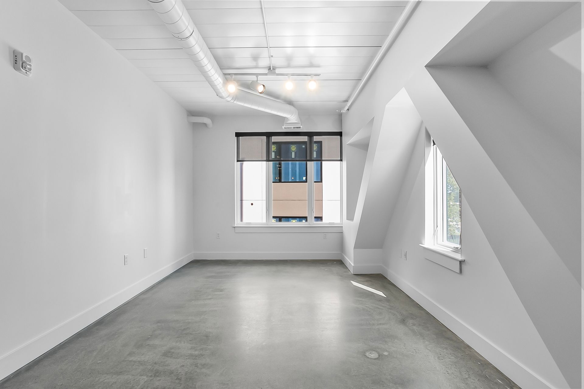 Empty, modern, white-walled room with concrete floor, window, and angled ceiling.