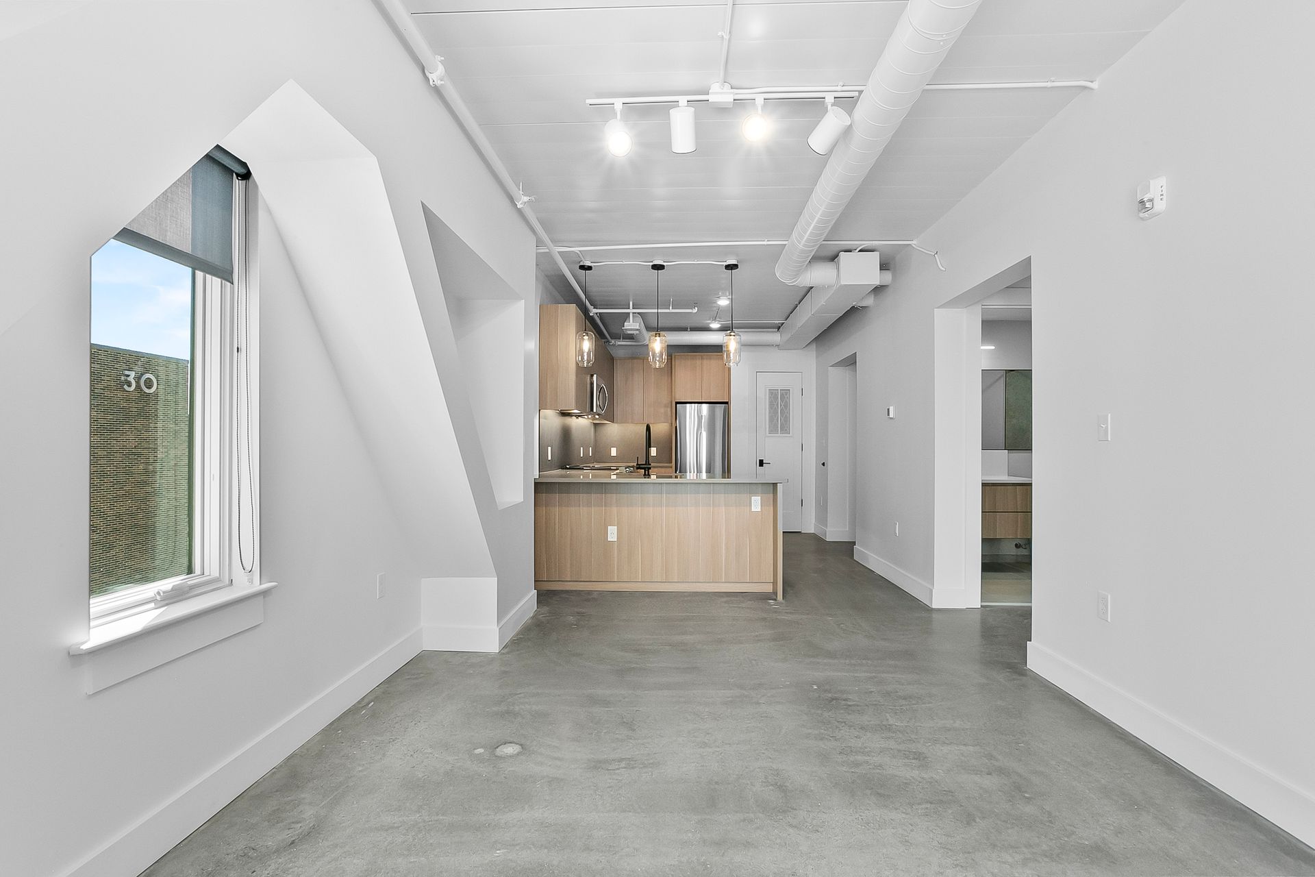 Empty, modern loft apartment interior with exposed ductwork, concrete floor, and kitchen in the background.