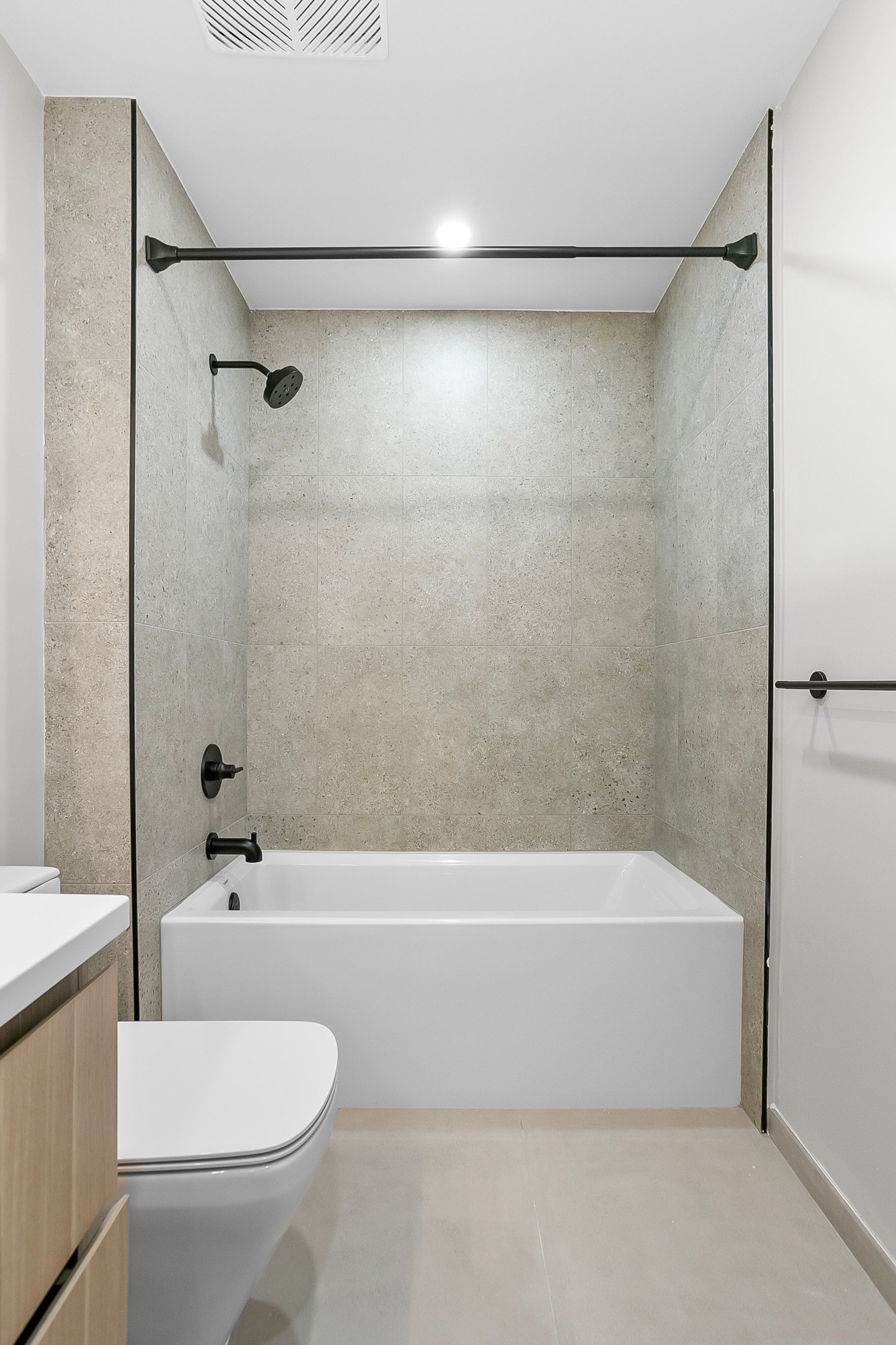 Bathroom with white tub, speckled shower wall, black fixtures, and light wood vanity.