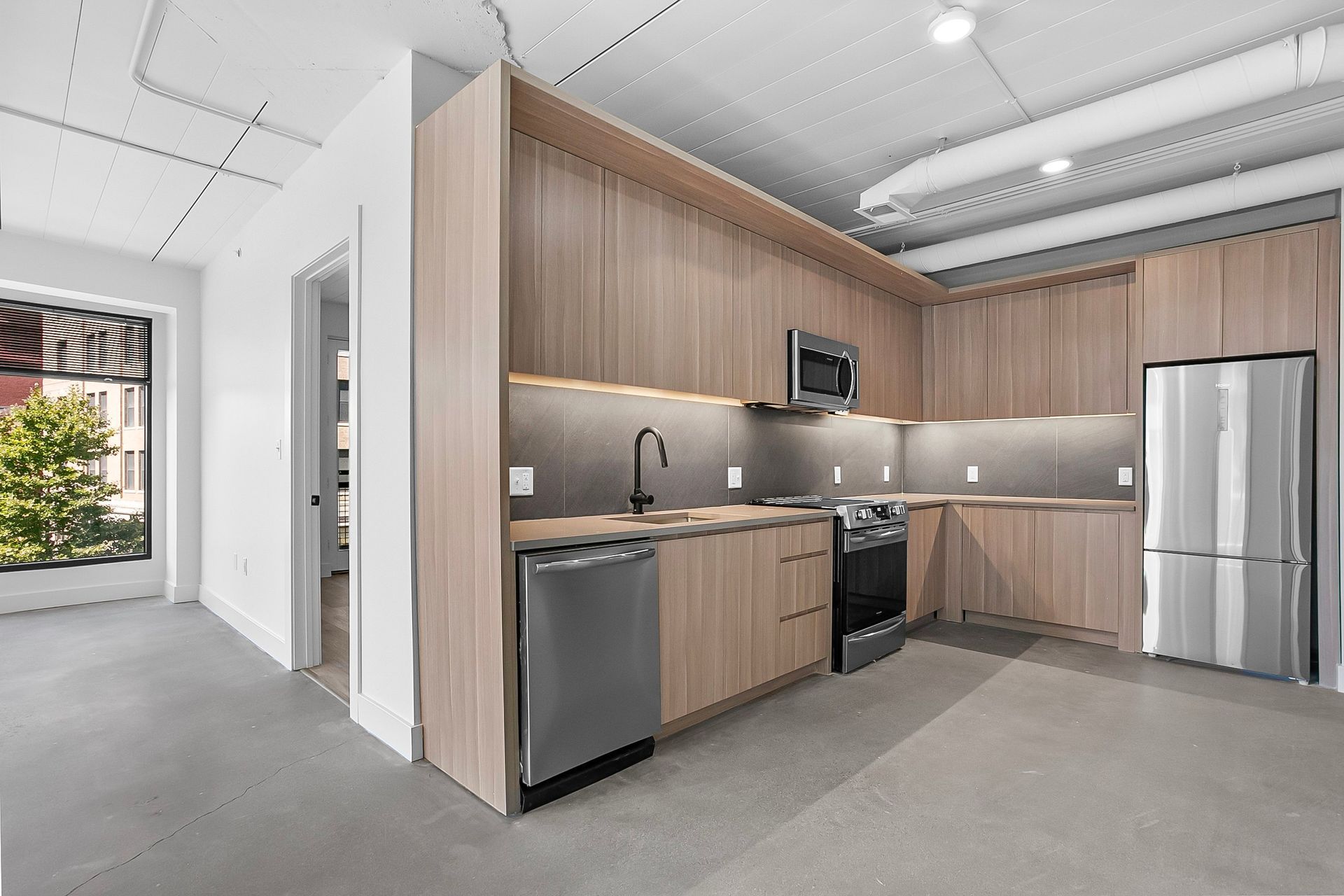 Modern kitchen with light wood cabinetry, stainless steel appliances, and concrete floor.