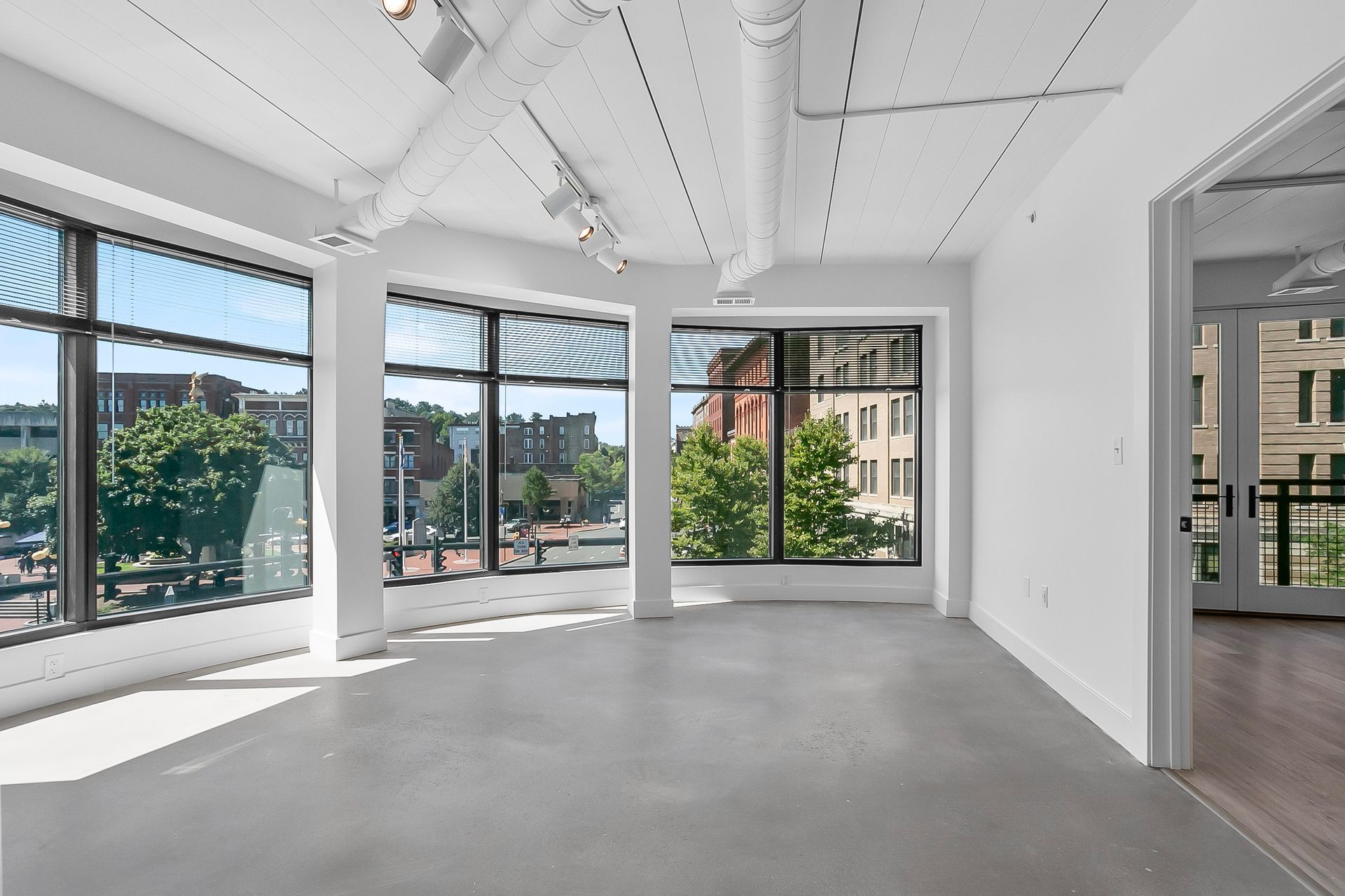 Empty room with large windows overlooking a city street. White walls, grey floor, natural light.