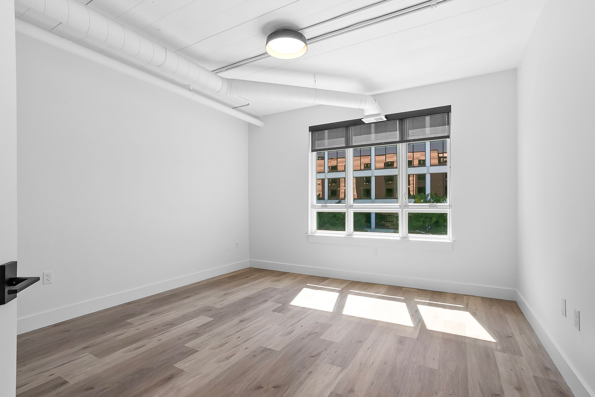 Empty room with light wood floor, white walls, window with blinds, and exposed ceiling ductwork.