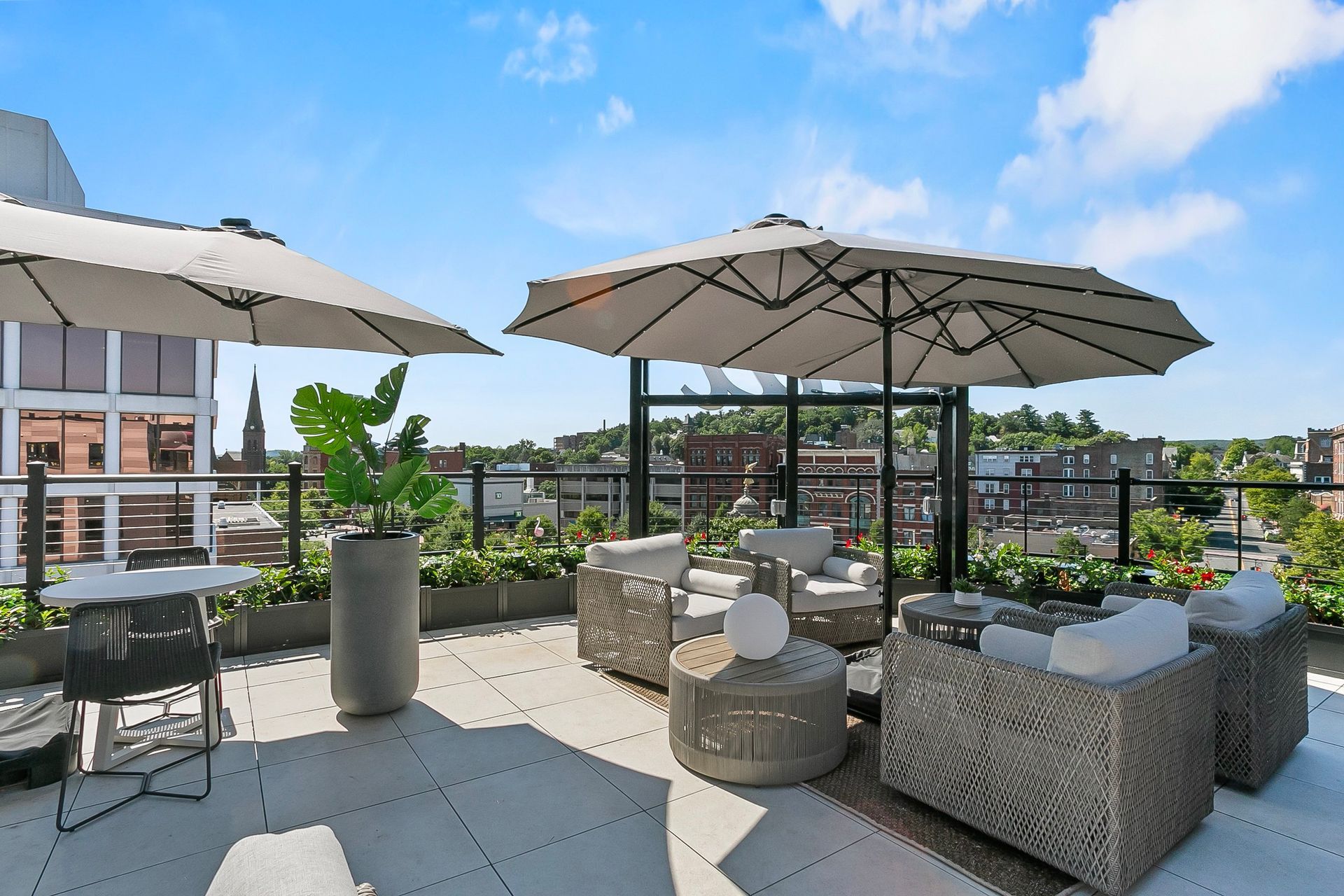 Rooftop patio with seating, umbrellas, and potted plants, overlooking cityscape on a sunny day.