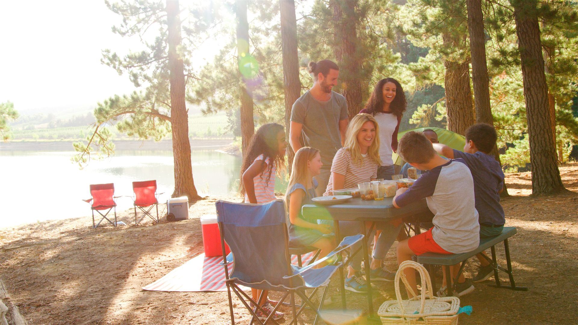 Friends gathered around a picnic table in a sunny lakeside park with red chairs and trees.