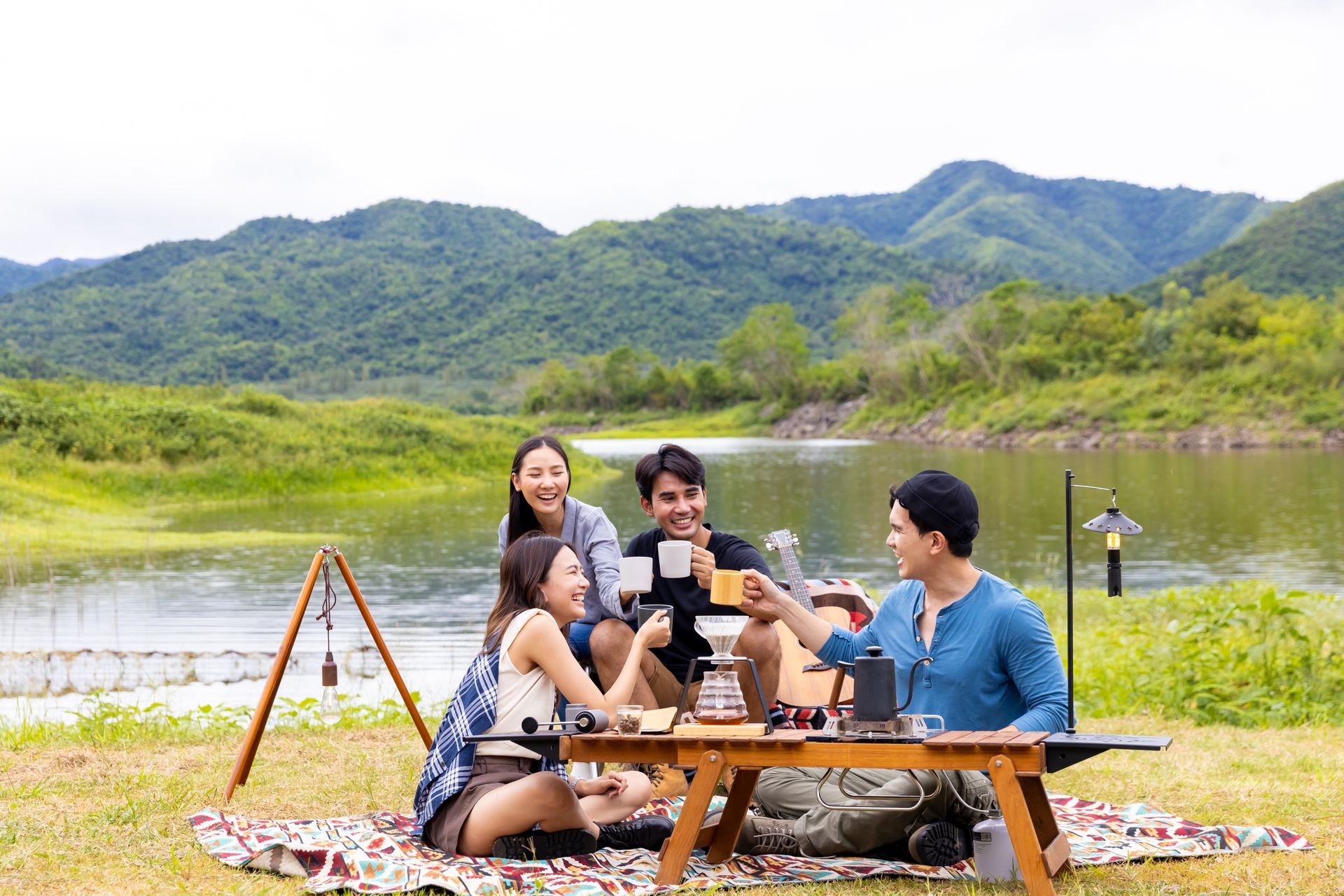 Group picnicking on a lakeside picnic mat with mountains and greenery in the background.