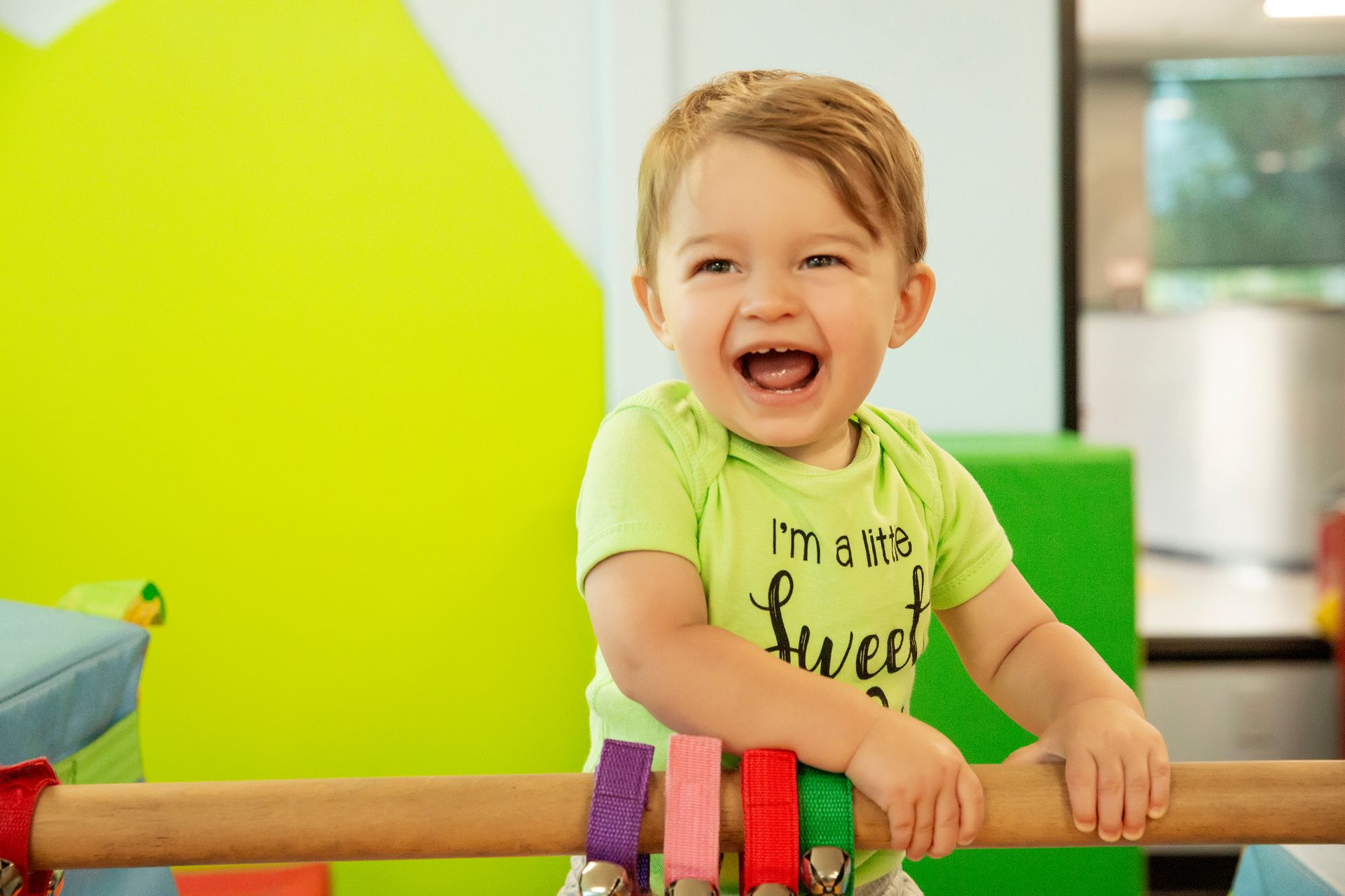 Smiling toddler in green shirt holding a wooden bar with colorful rings, in a brightly colored playroom.